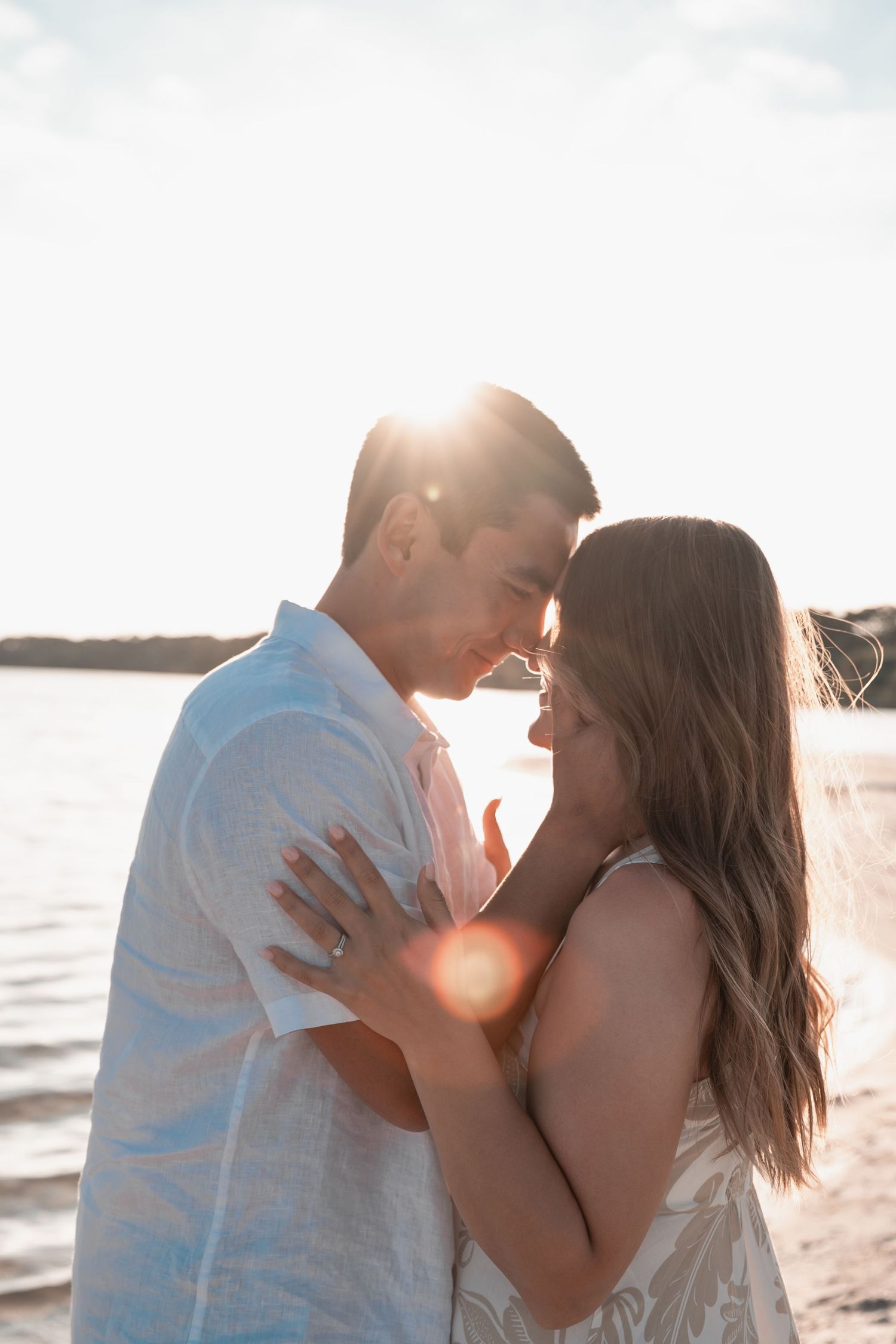 Couple embracing on a beach at golden sunset with warm lens flare, romantic engagement photo by the water.