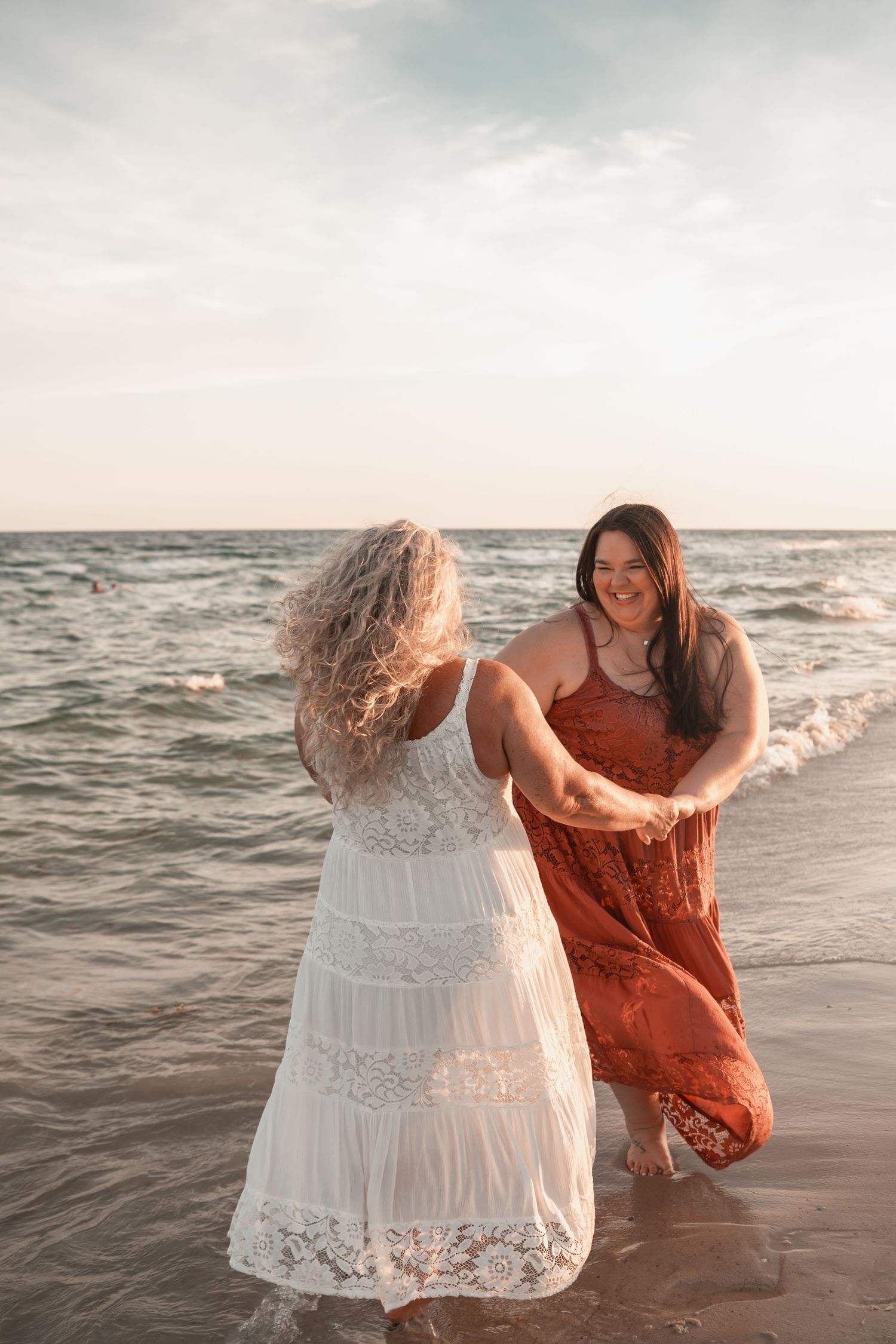 Two women in flowing dresses dancing on a beach at sunset, one in white lace and one in rust red.