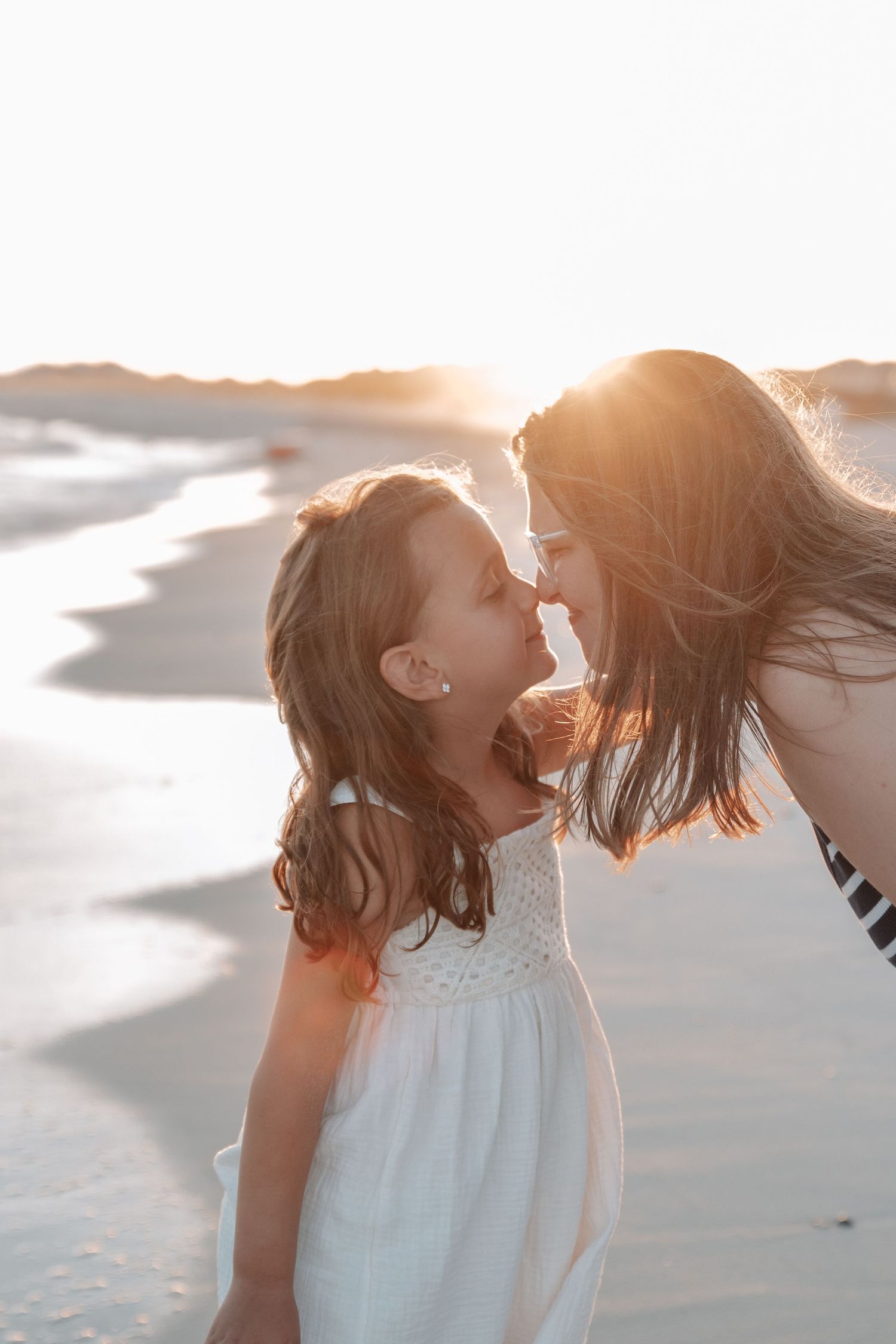 A mother and young daughter share a tender nose-to-nose moment on a sunlit beach at golden hour.
