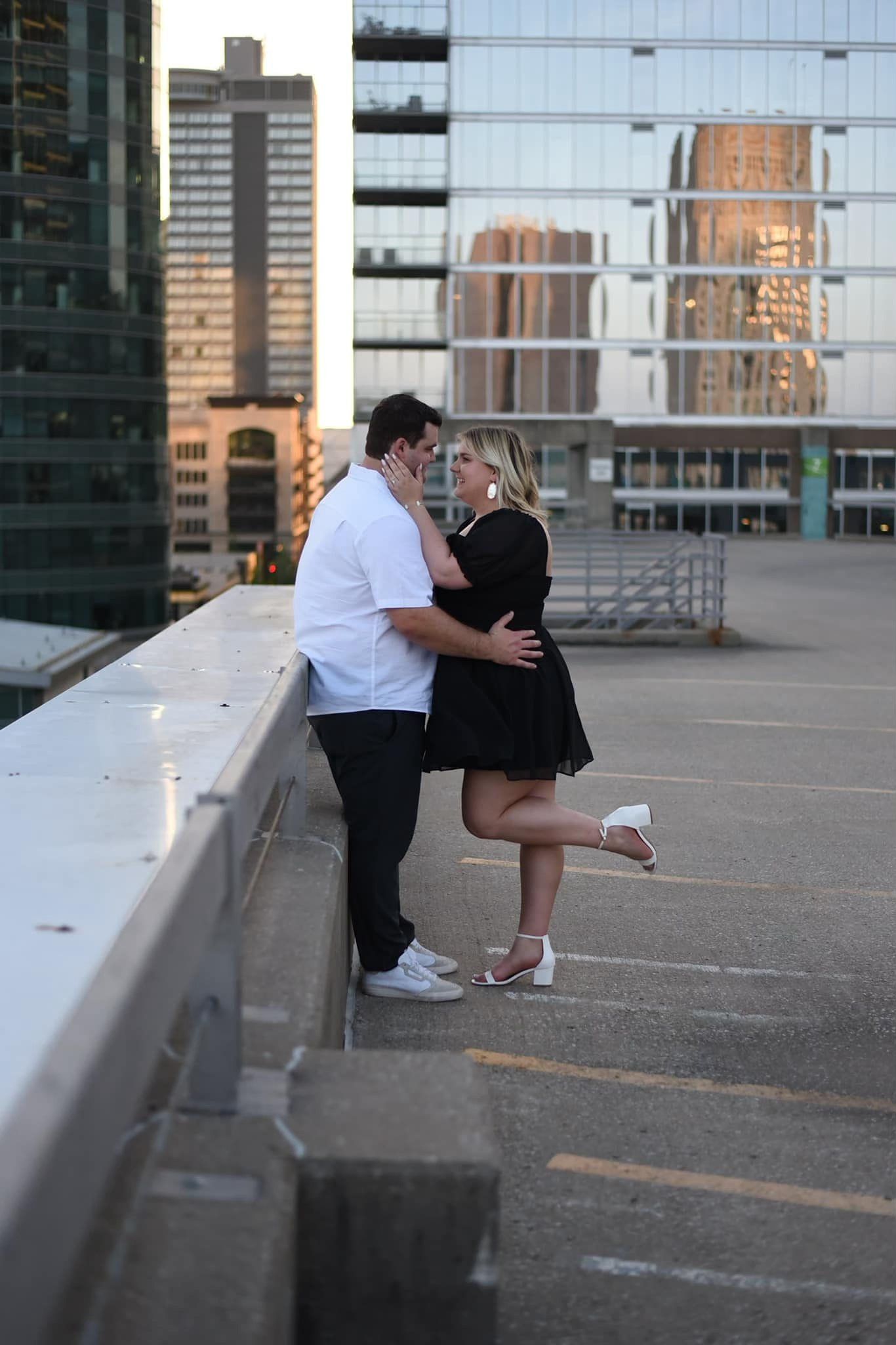 Couple sharing a romantic kiss on a rooftop parking garage with city skyline buildings reflected in glass behind them.
