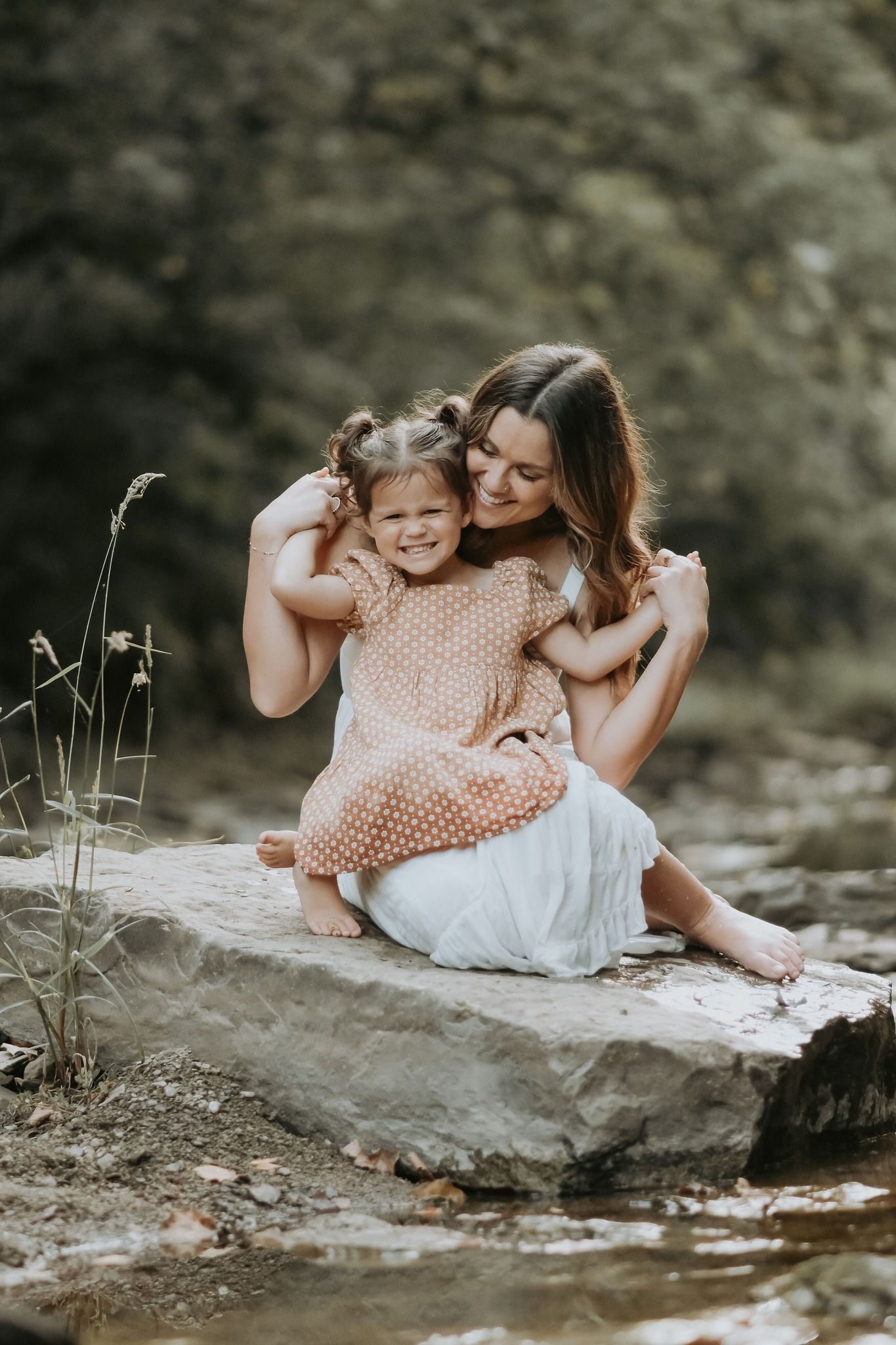 A mother sits on a rock by a creek hugging her laughing toddler daughter in a pink dress.