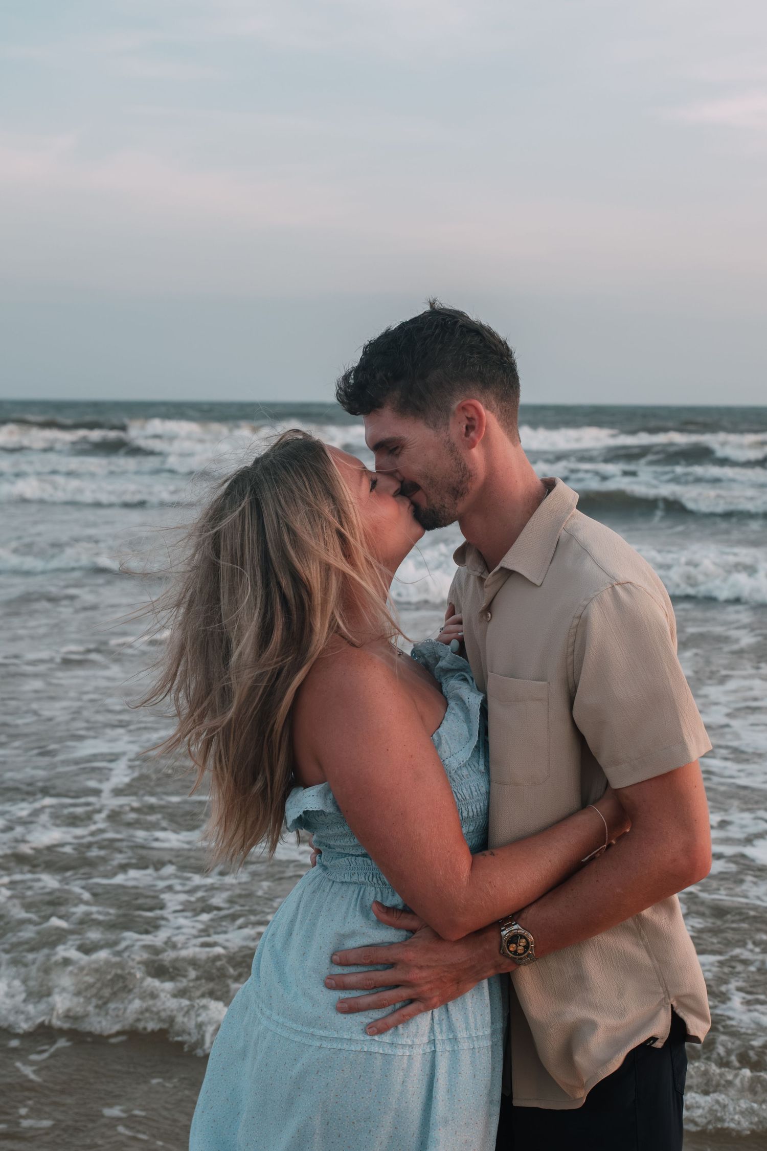 A couple shares a romantic kiss on a sandy beach at sunset, with ocean waves crashing behind them.