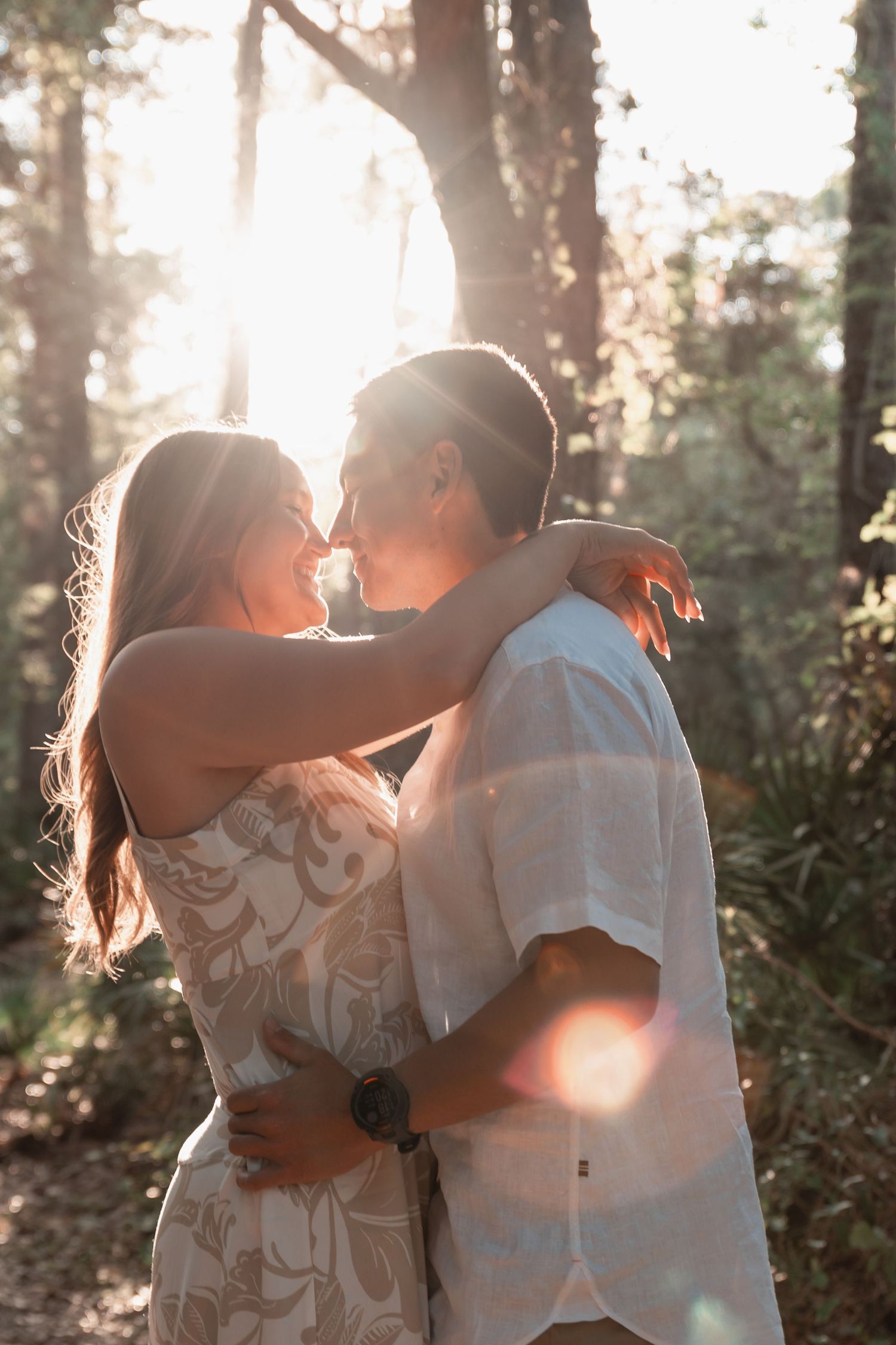 Couple embraces nose-to-nose in a glowing sunlit forest, warm lens flare highlights their romantic moment.