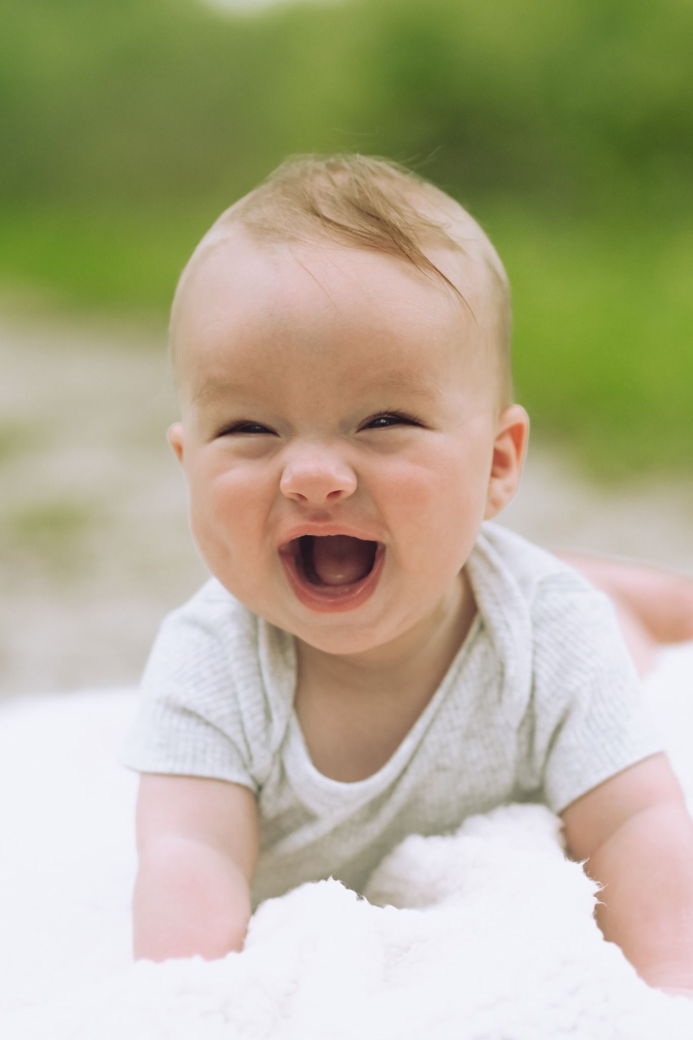 An adorable laughing baby lies on a white blanket outdoors on a sunny day.