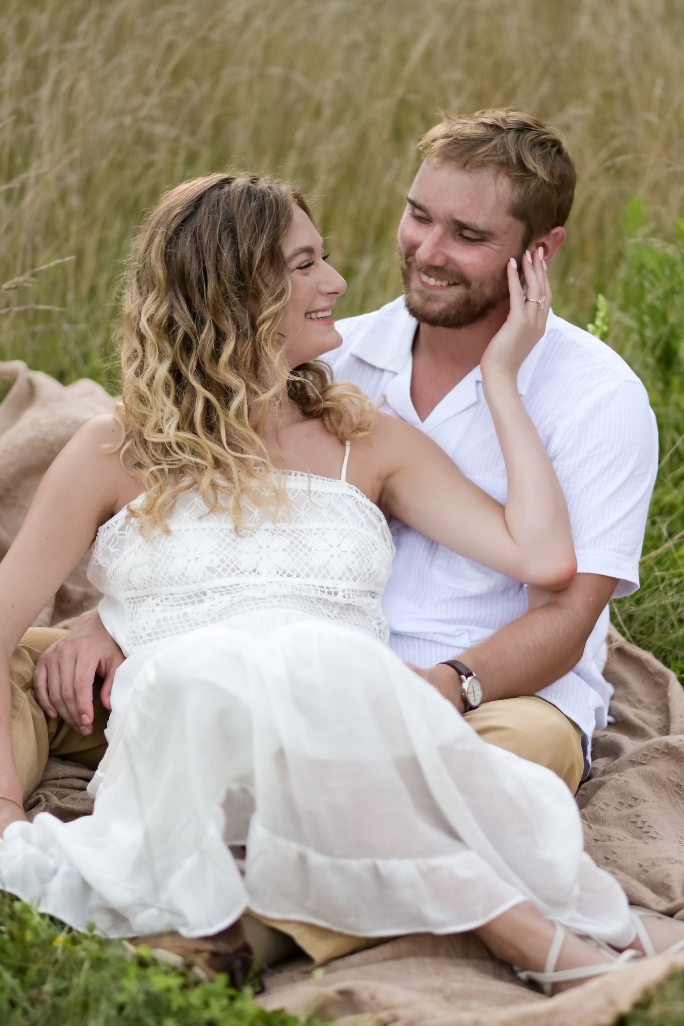 Smiling couple sitting together on a blanket in a golden grass field, sharing a tender romantic moment.
