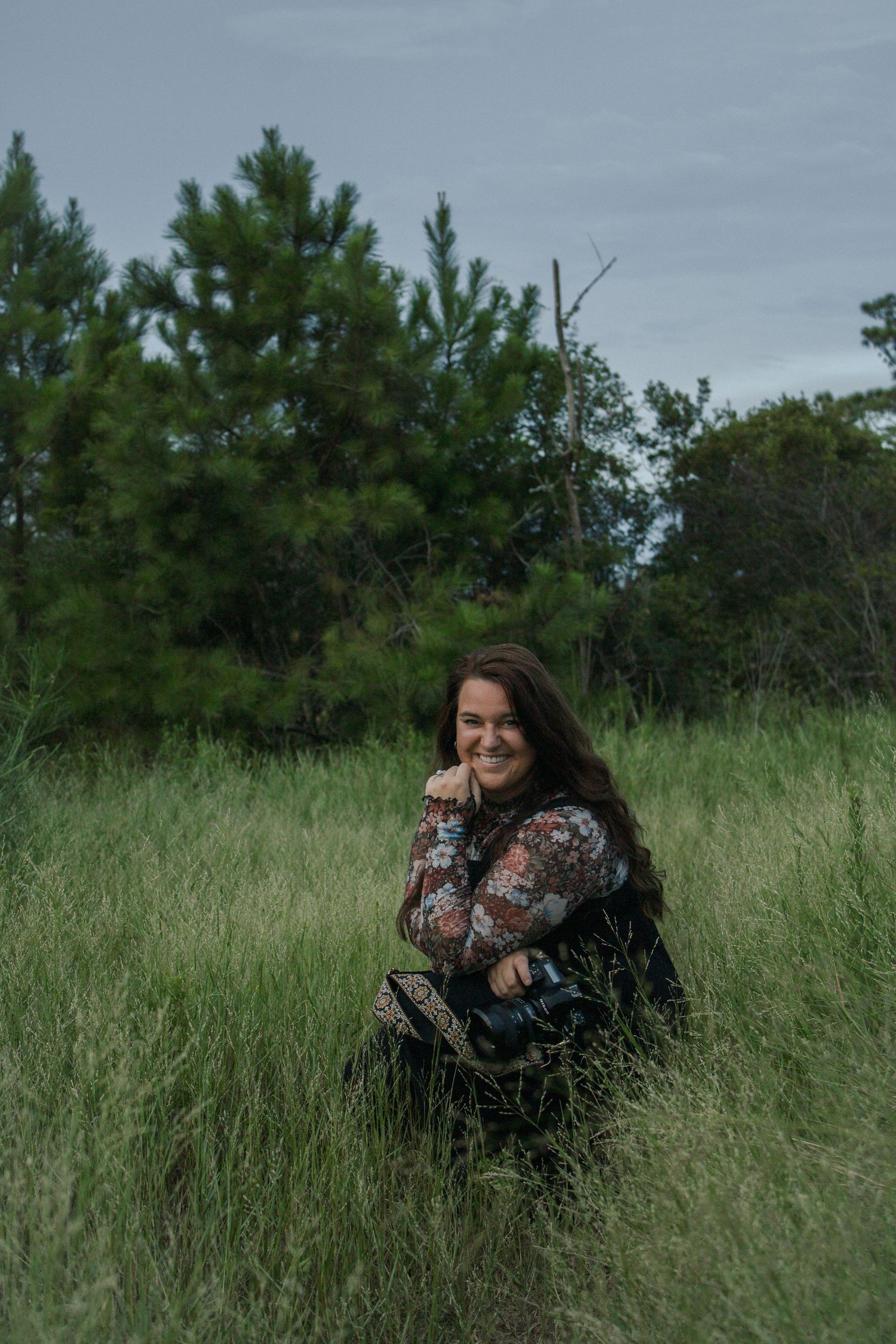 A smiling woman with long brown hair sits in tall green grass, wearing a floral top, with pine trees behind her.