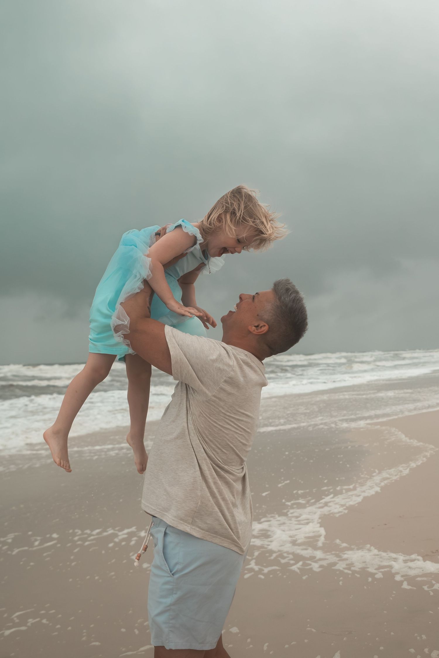 A father joyfully lifts his young daughter in a blue dress on a cloudy beach with ocean waves behind them.