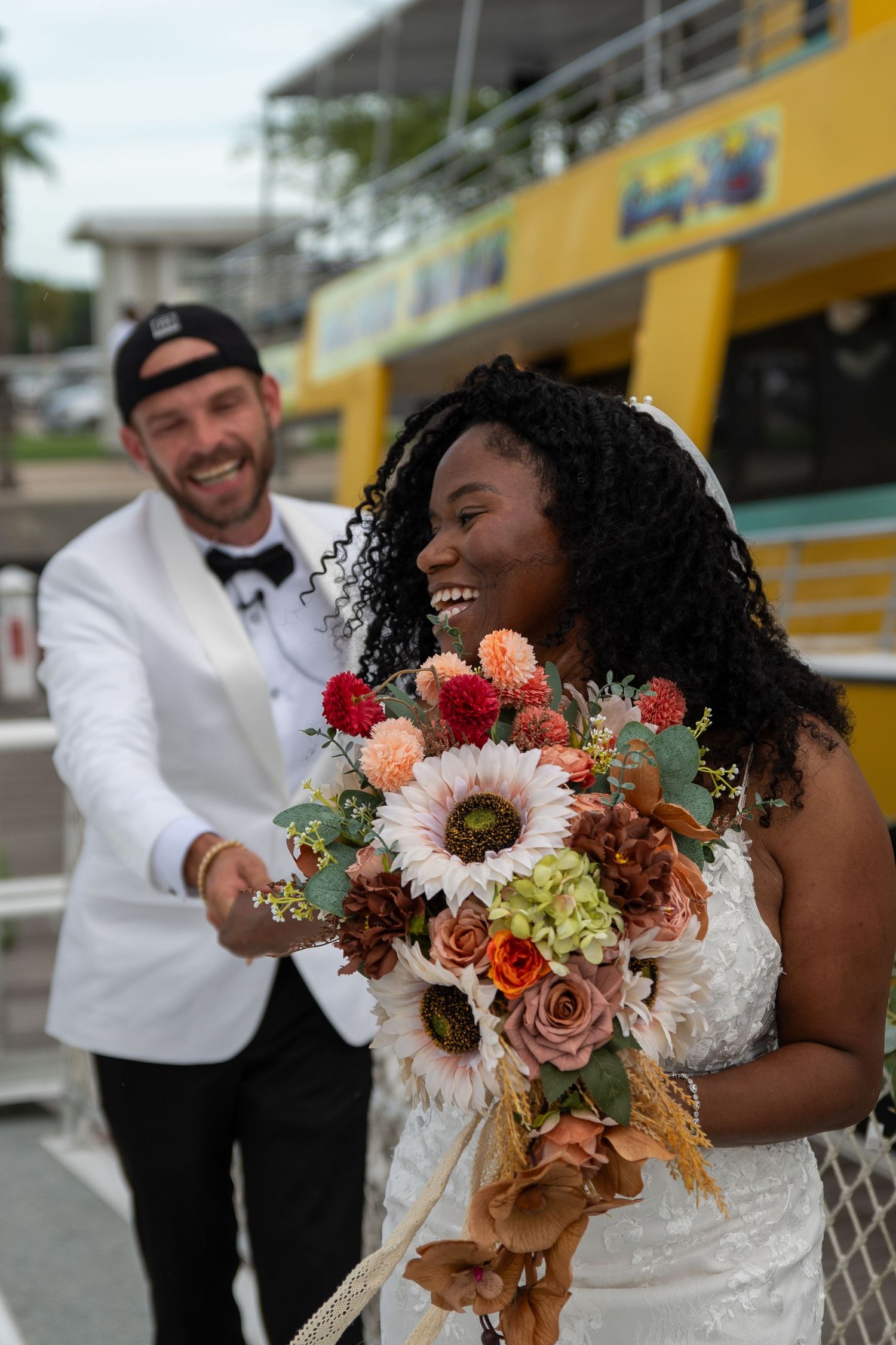 Interracial couple in wedding attire, bride holding colorful bouquet with sunflowers and roses, groom in white tuxedo smiling.