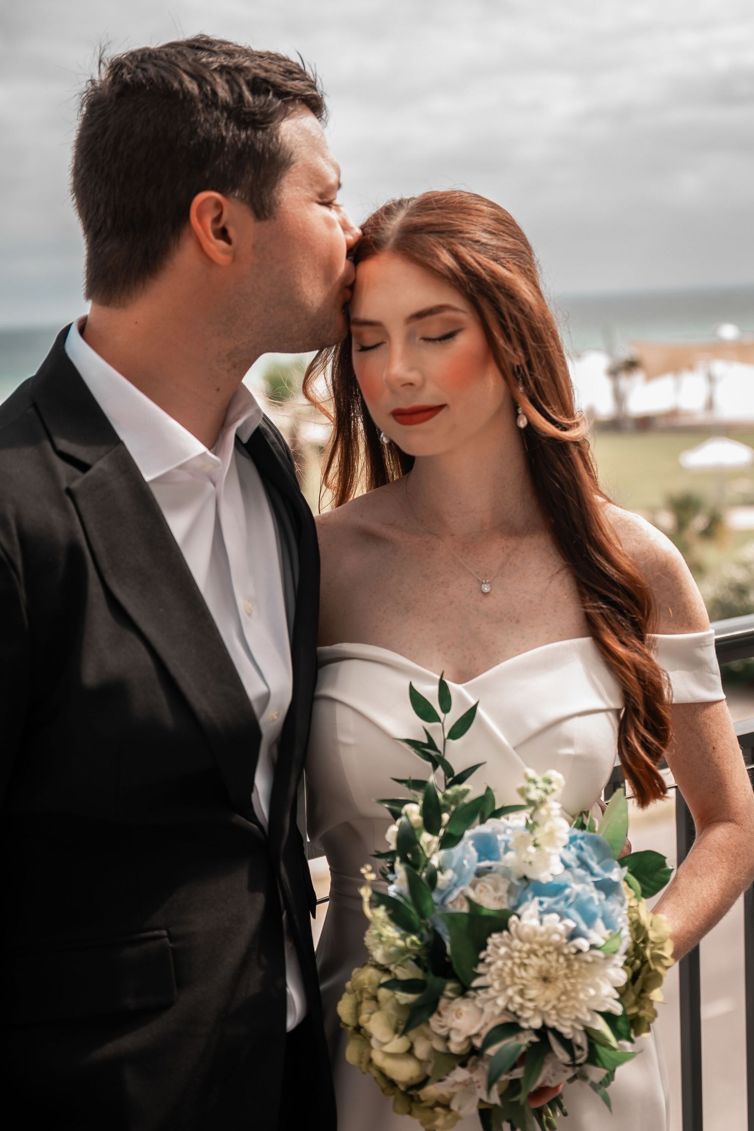 Groom in black suit kissing bride with red hair holding a blue and white bouquet on balcony.