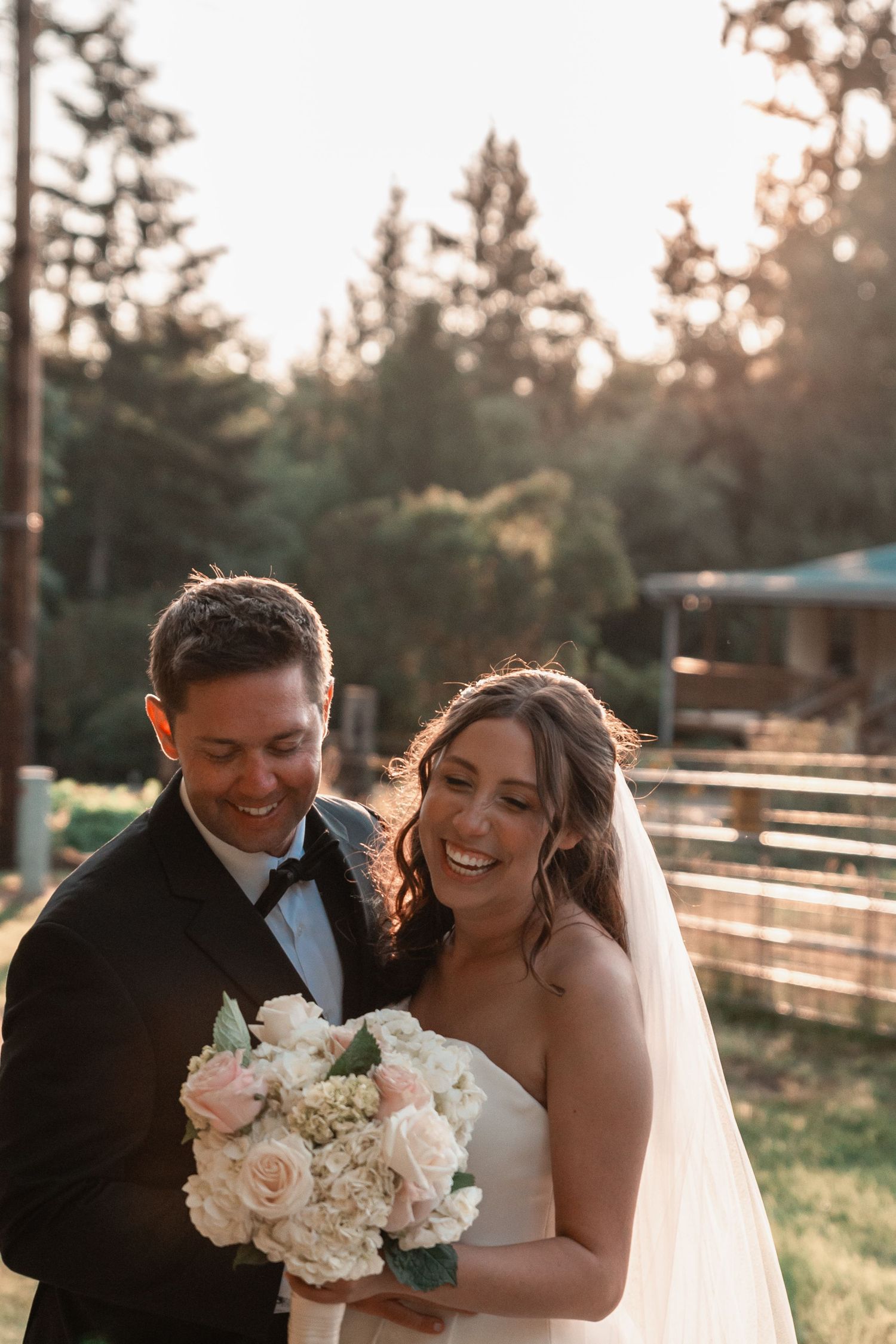 Smiling bride and groom in wedding attire holding white floral bouquet outdoors with golden sunset light and trees behind them.