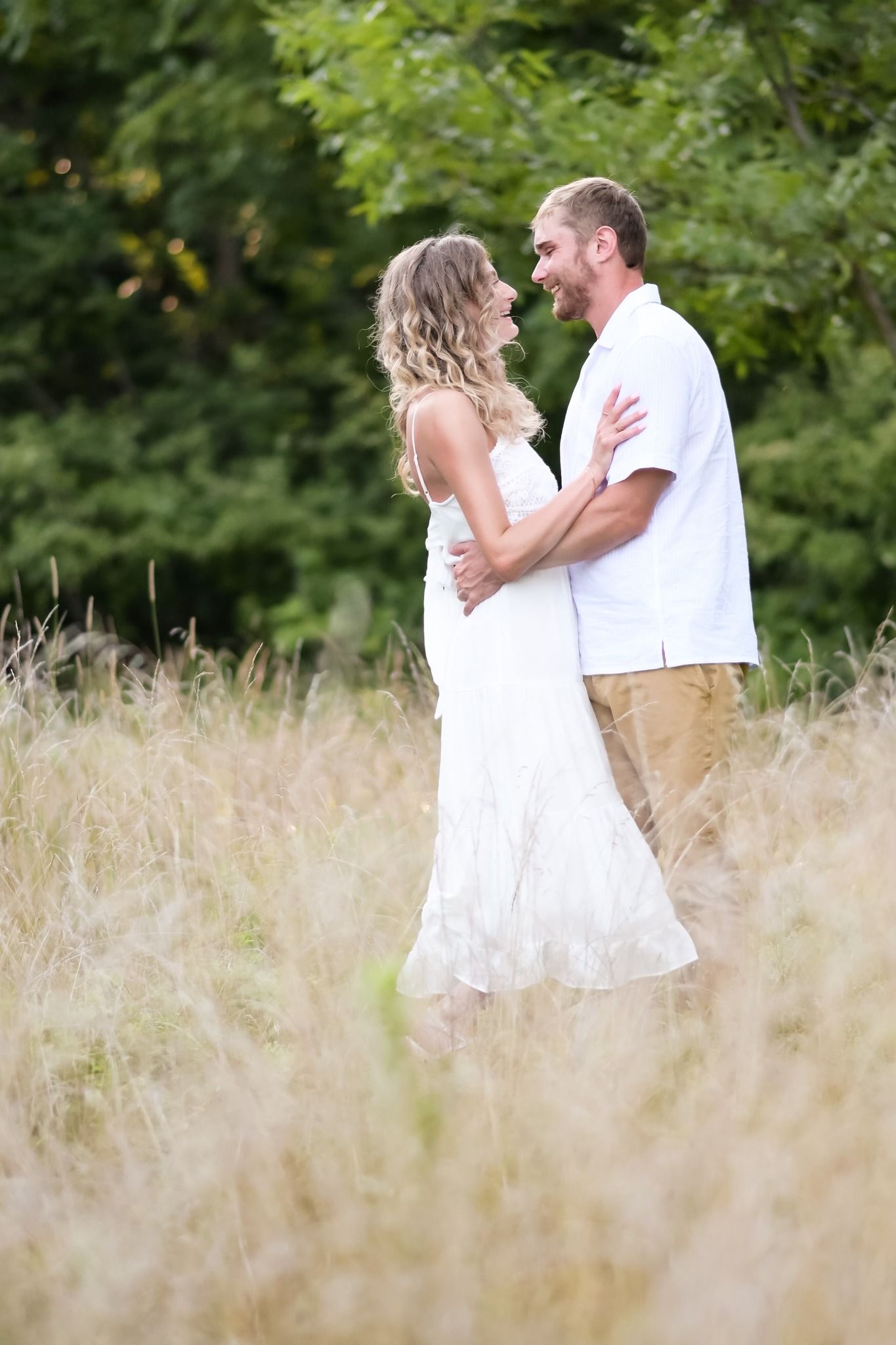 Couple embracing and smiling in a field of tall golden grass with green trees in the background.