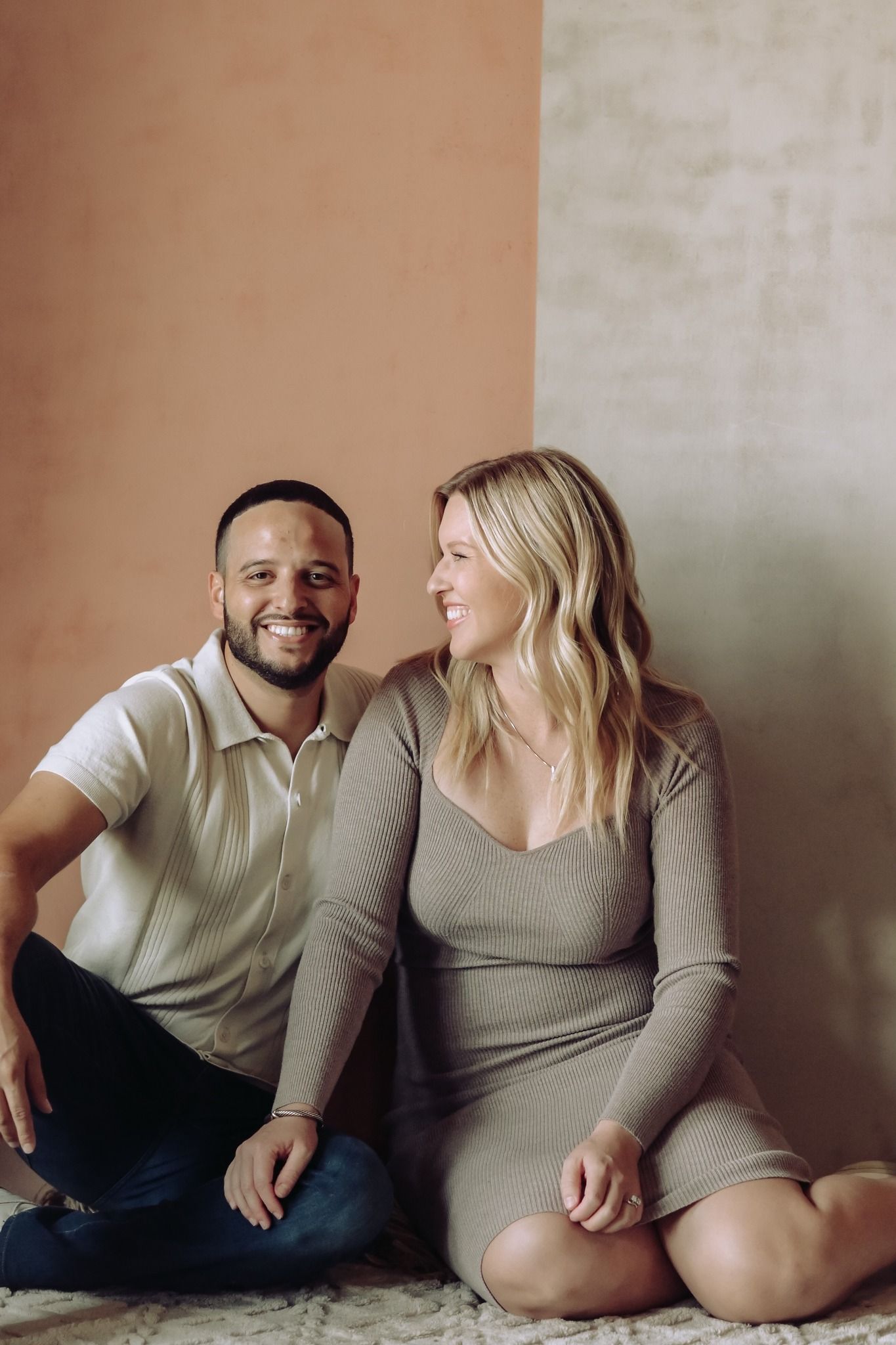Smiling couple sitting on the floor against a warm terracotta wall in a cozy indoor setting.