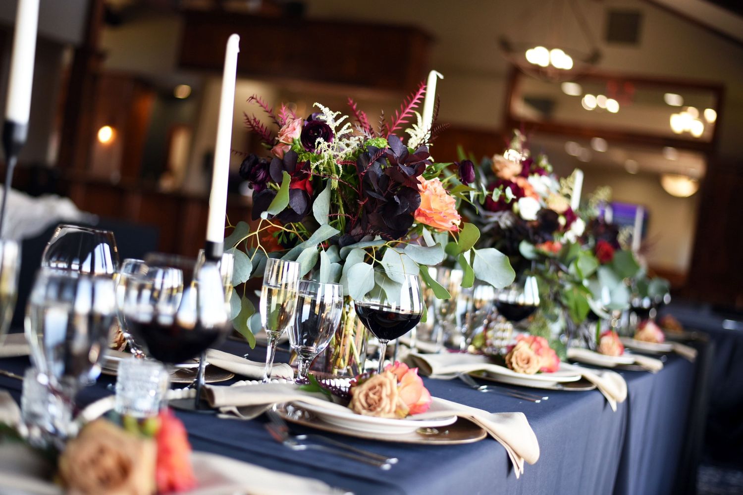 Elegant wedding reception table with dark floral centerpiece, navy tablecloth, candles, and wine glasses in ballroom.