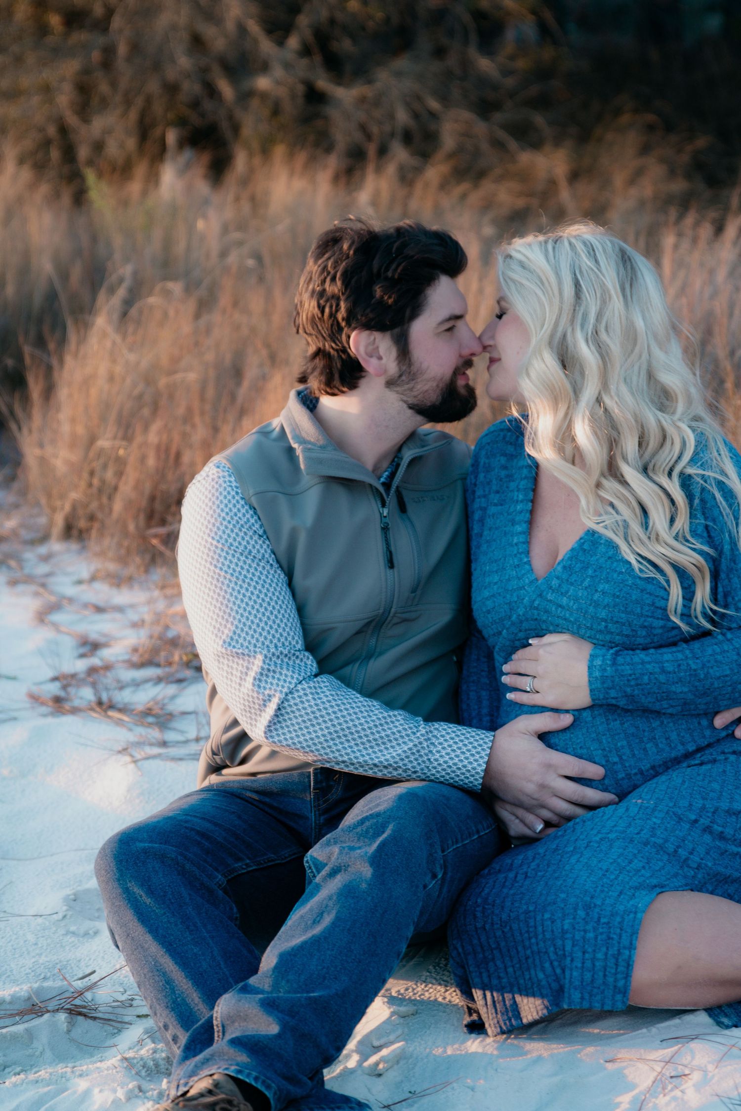 Expecting couple shares a tender kiss while cradling baby bump on sandy outdoor ground.