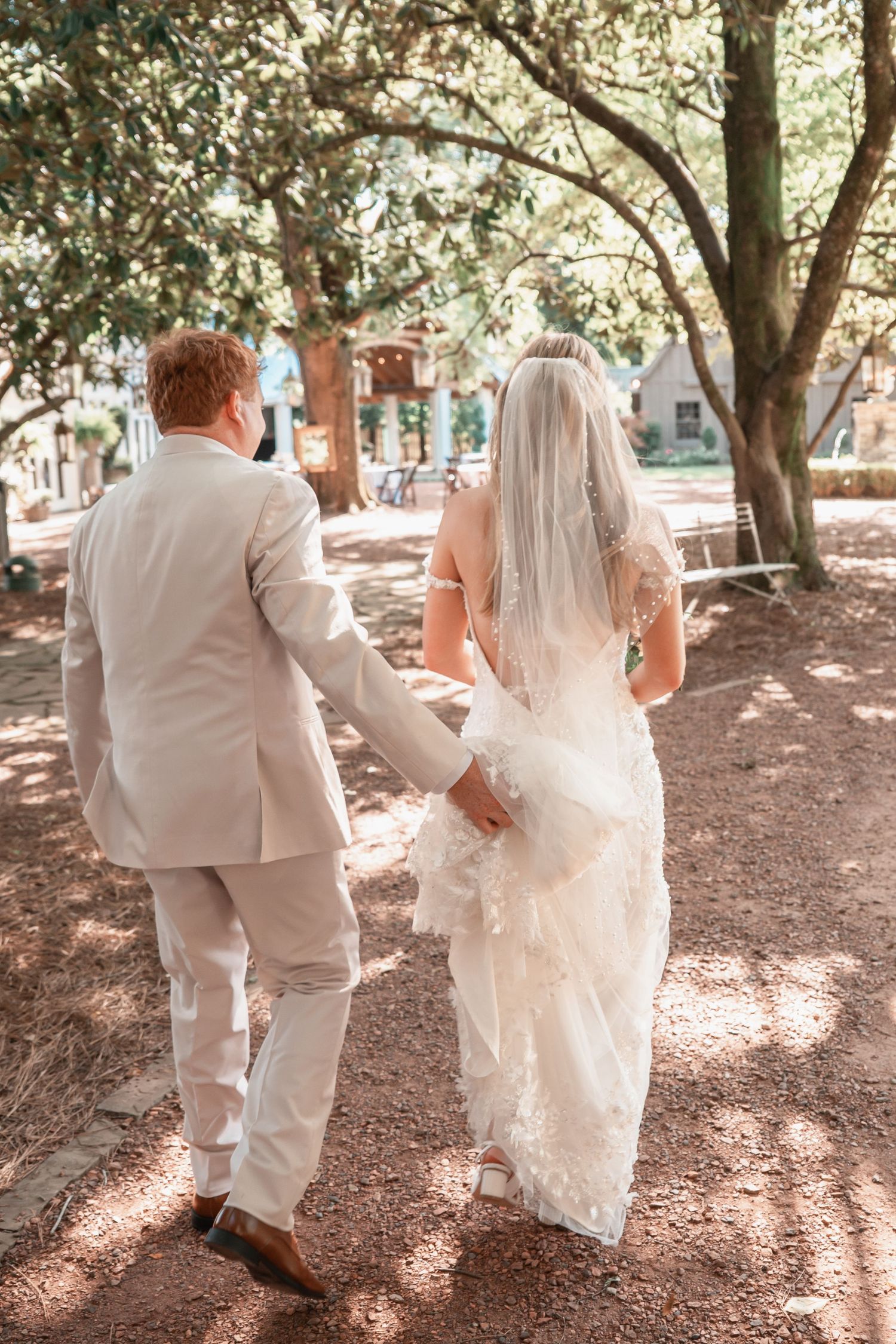 A bride and groom walk hand-in-hand through a tree-lined outdoor venue, she in a lace gown with veil, he in a tan suit.