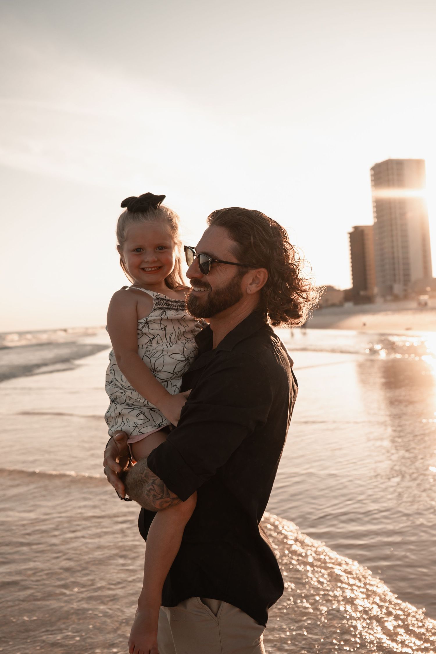 Bearded father holding young daughter at golden sunset on a beach with city skyline.
