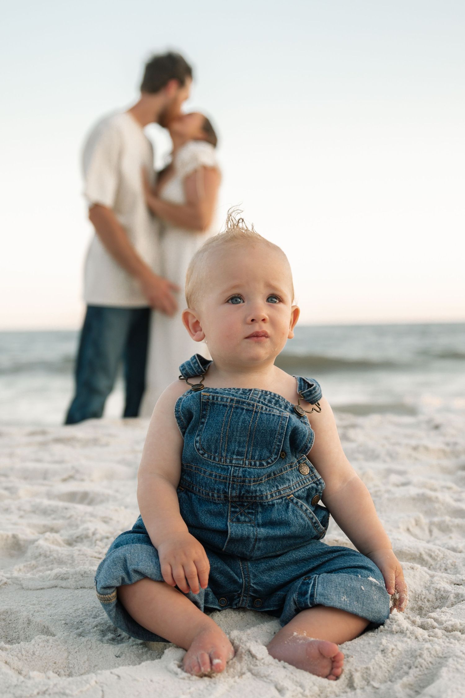 Baby in denim overalls sits on sandy beach while parents share a romantic moment by the ocean at sunset.