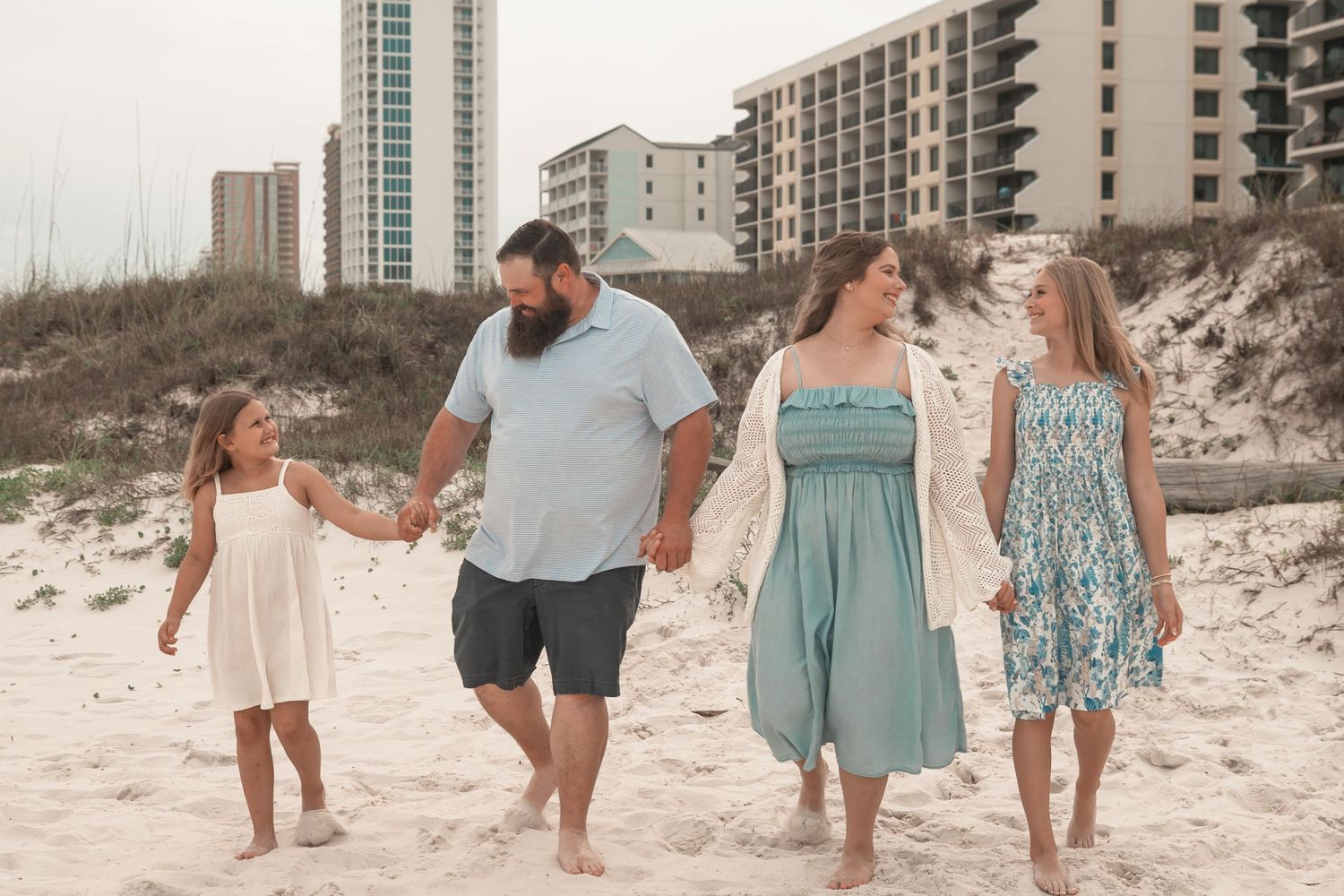 Family of four walking hand-in-hand on a sunny beach with sand dunes and coastal buildings in the background.