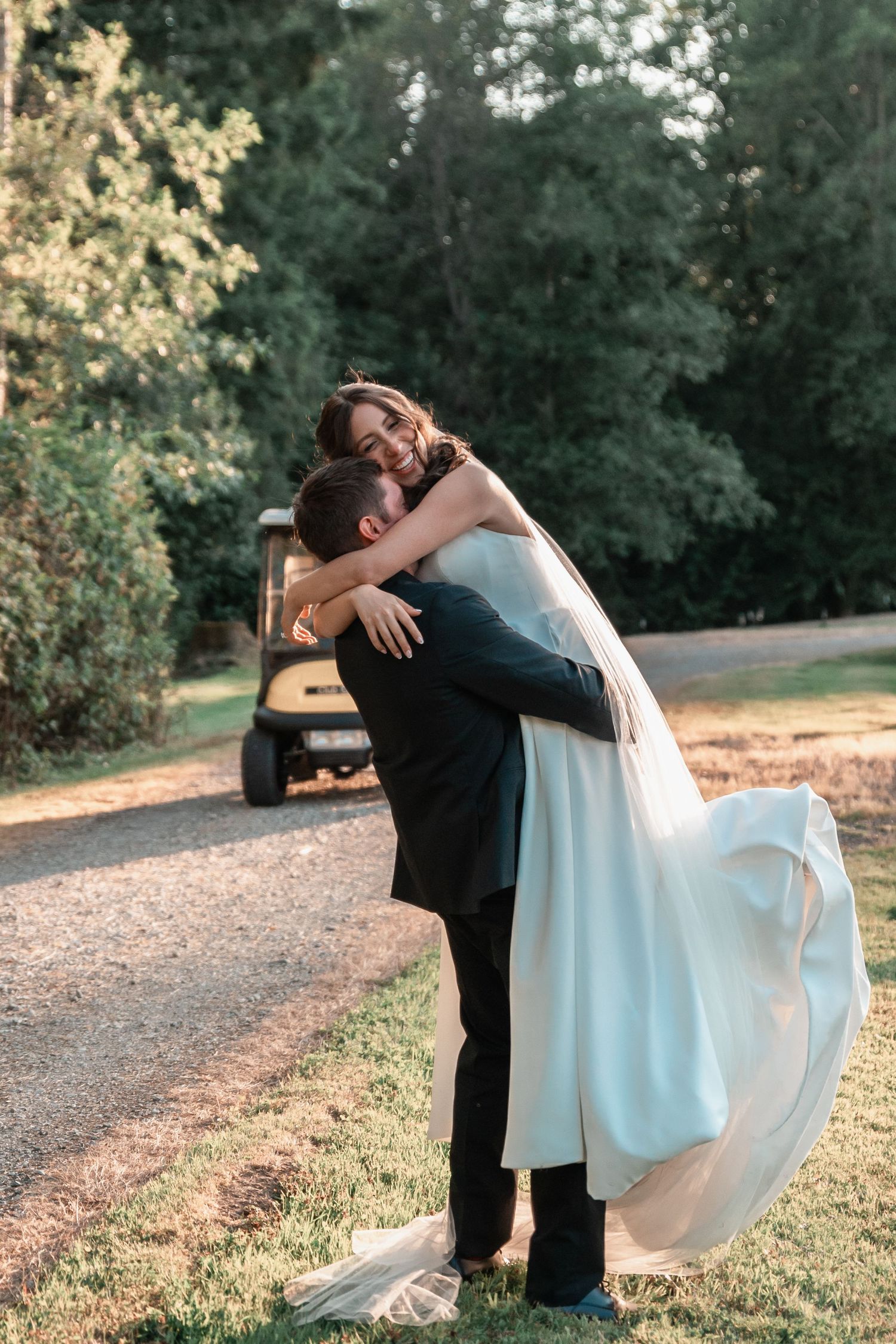 Groom lifts bride in white gown outdoors, sharing a joyful embrace on a sunlit path surrounded by lush green trees.