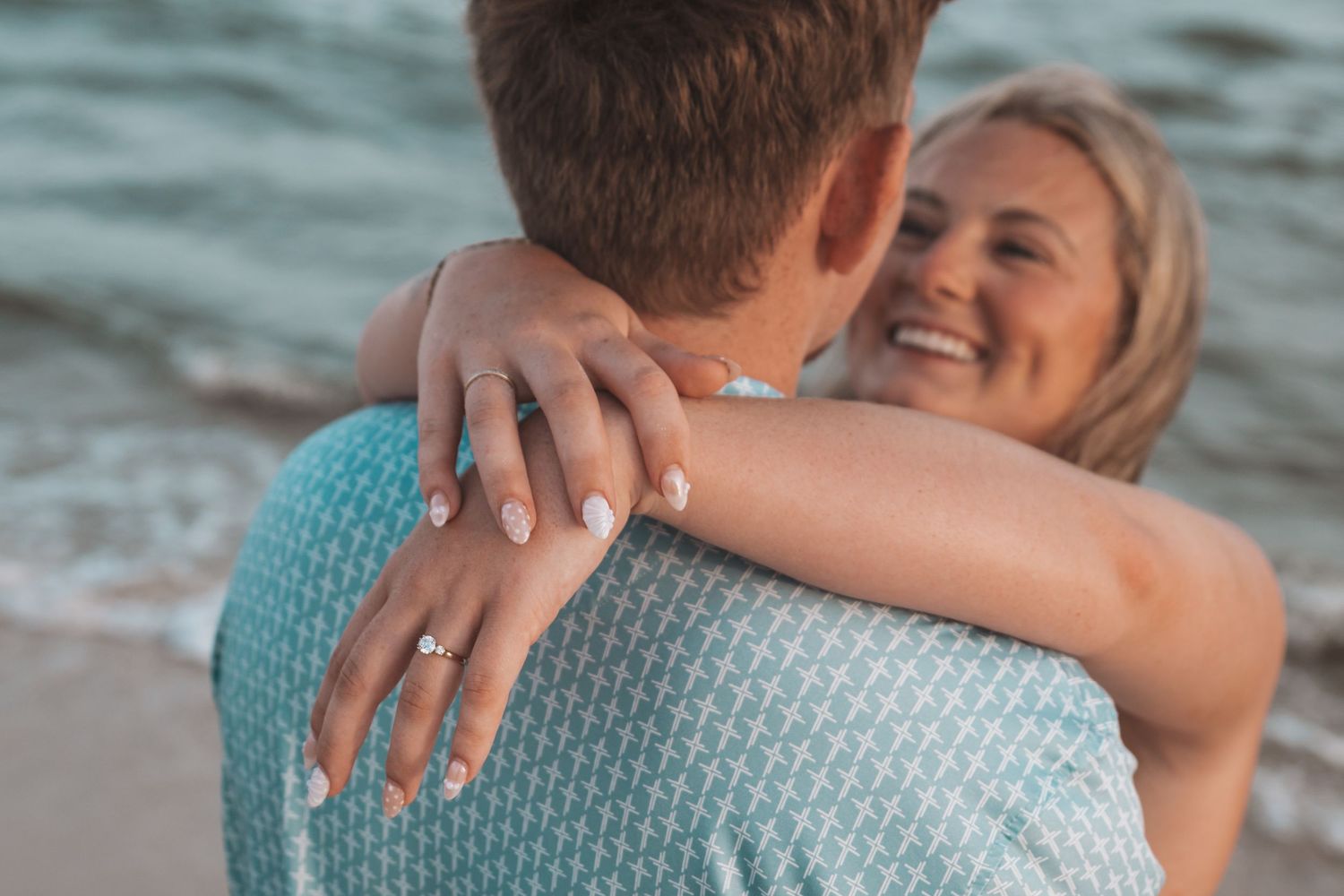 Couple embracing on beach, woman smiling with engagement ring visible on hand, ocean waves in background.