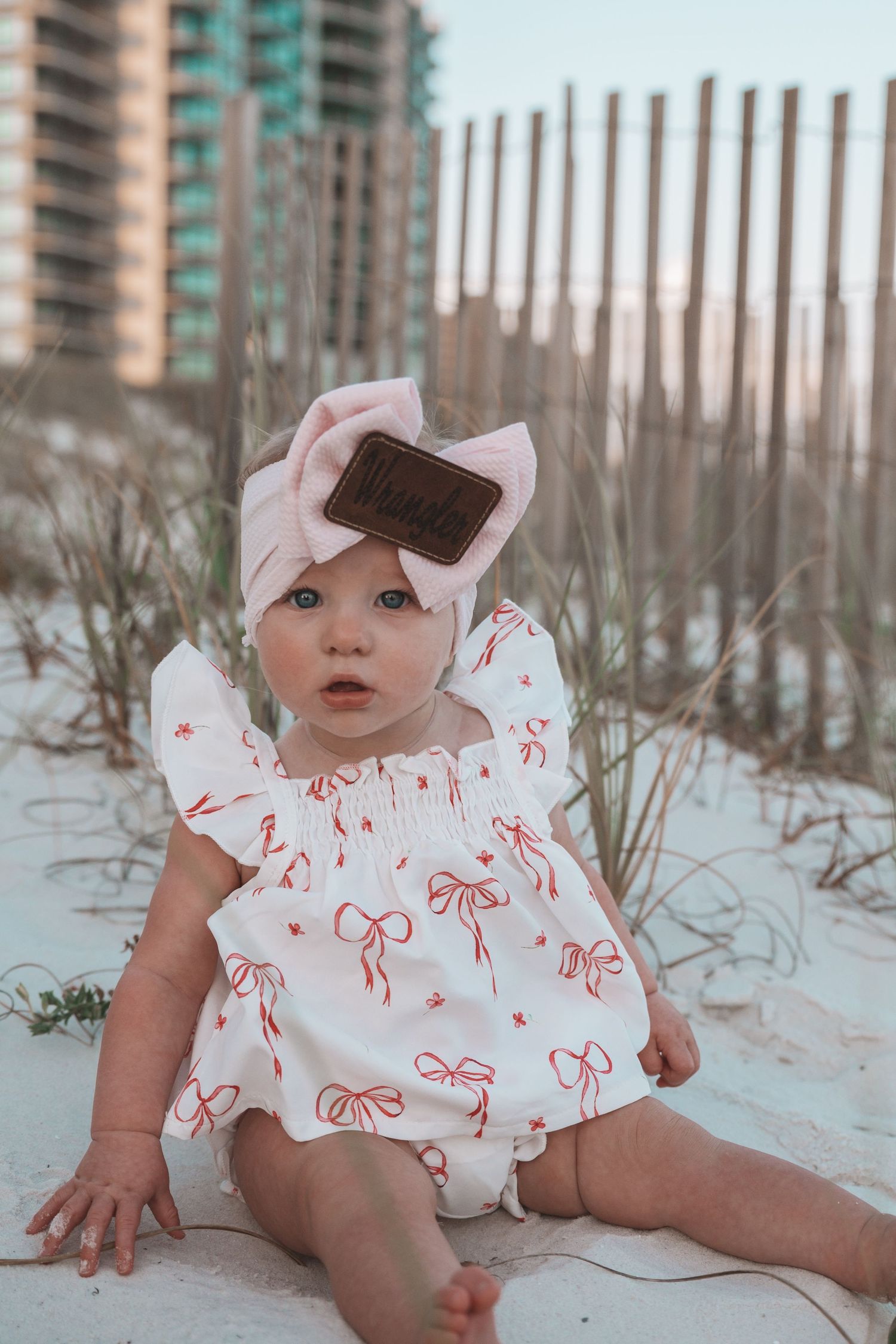 Baby girl in white bow-print romper and pink headband sits on white sand beach with dunes and buildings in background.