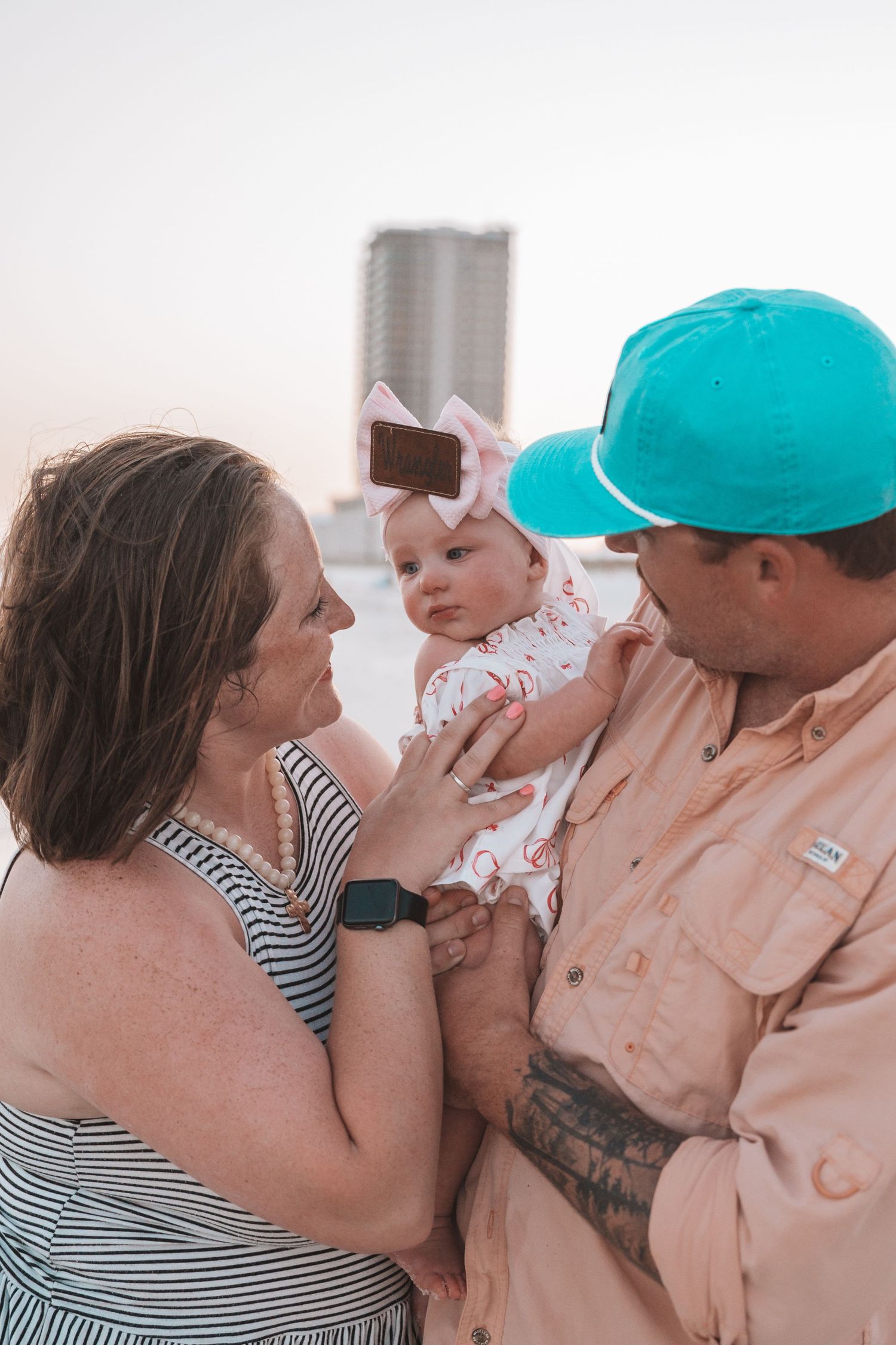 A couple lovingly holds their baby girl wearing a pink bow on a beach with a city skyline backdrop.
