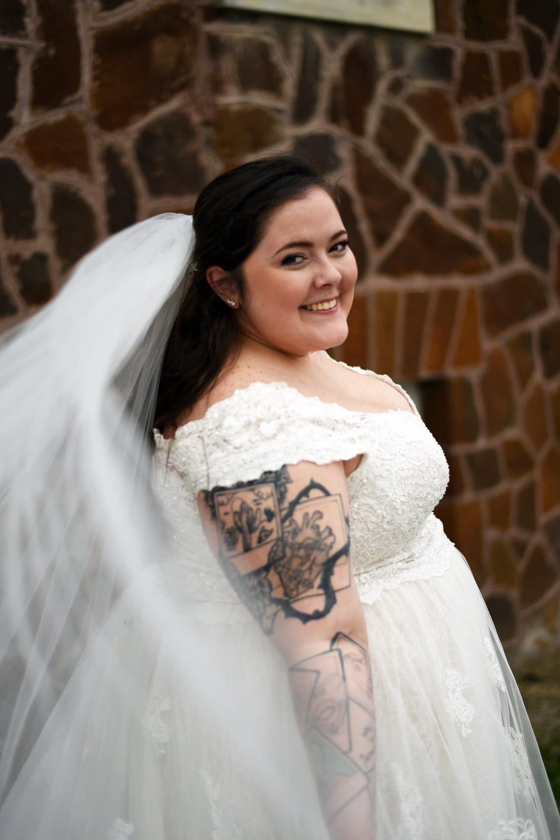 Plus-size bride in off-shoulder lace wedding gown with sleeve tattoos smiling while holding flowing veil outdoors.