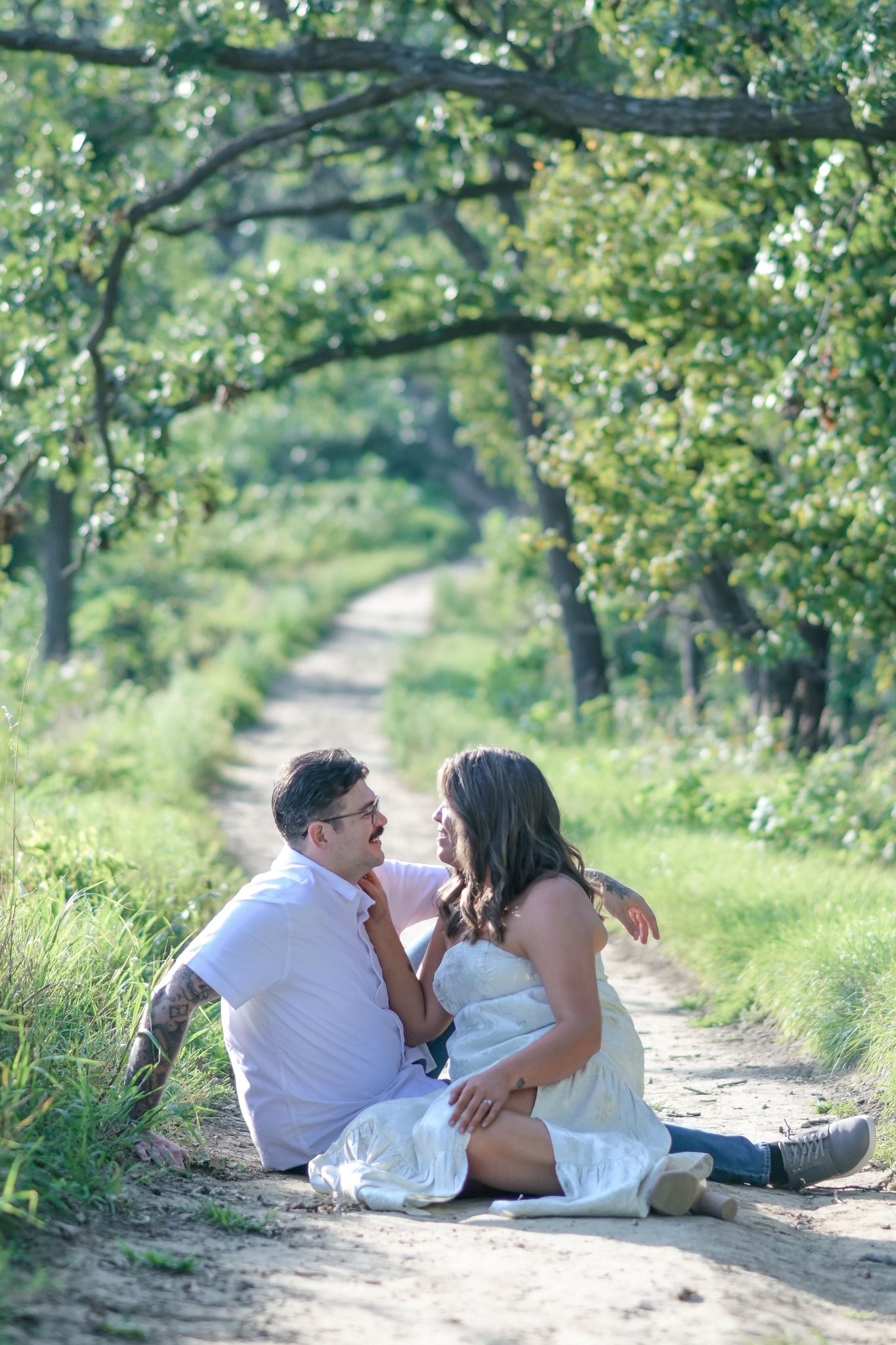 Couple sitting together on a scenic tree-lined dirt path, sharing an intimate moment outdoors.