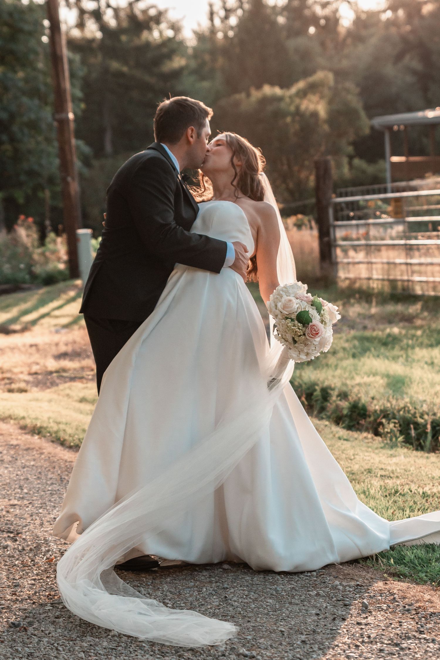 A bride and groom share a romantic kiss outdoors at golden hour, her white gown flowing gracefully as she holds a bouquet.