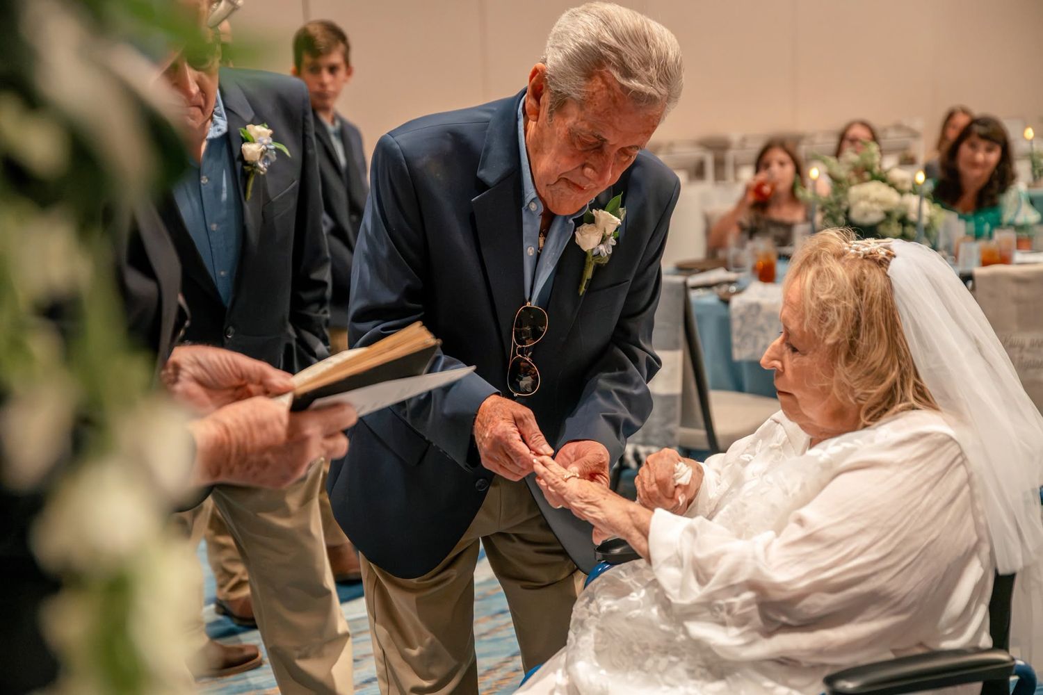Elderly couple exchanging rings during wedding ceremony, groom in navy suit, bride in white veil seated in wheelchair.