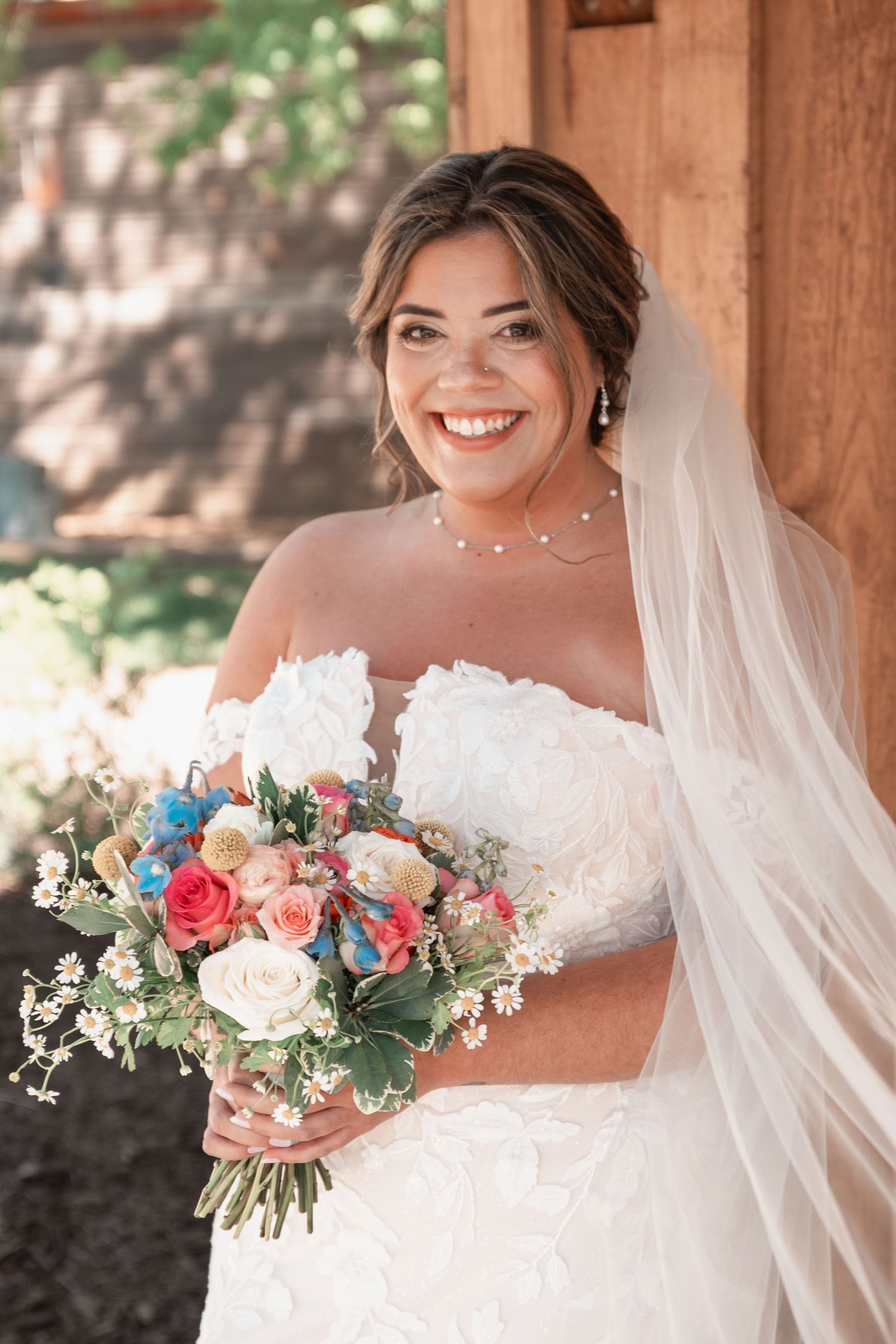Radiant bride in lace off-shoulder gown holding colorful bouquet with veil flowing at barn venue.