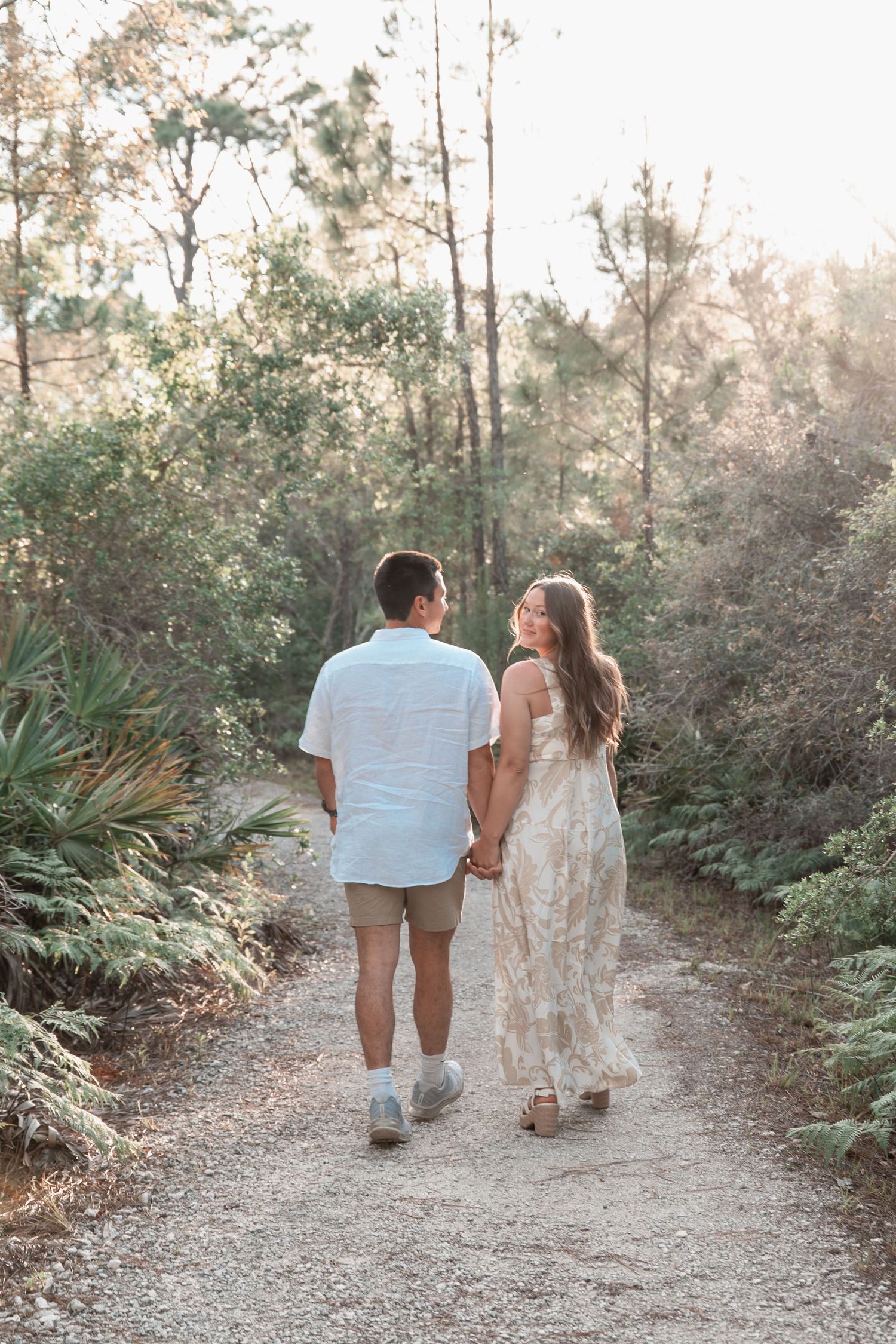 Couple walks hand-in-hand down a sunlit gravel path surrounded by lush green trees and tropical foliage.