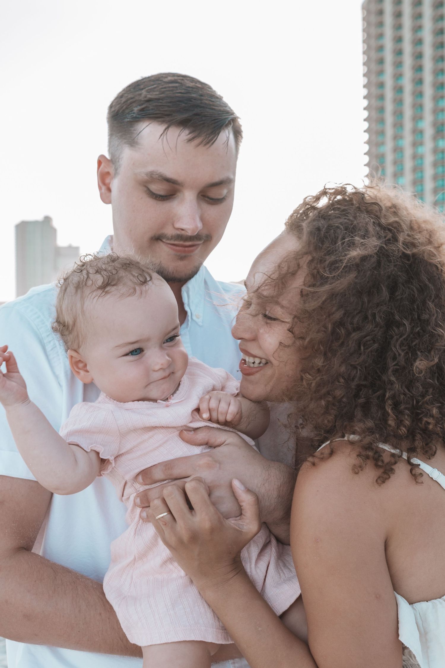 Happy young family with mother, father, and baby in pink outfit posing together outdoors with city skyline behind.