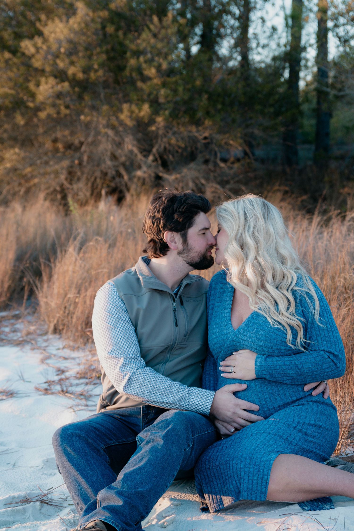 Couple sharing a tender kiss while sitting on snowy ground outdoors, woman in blue knit dress showing baby bump.