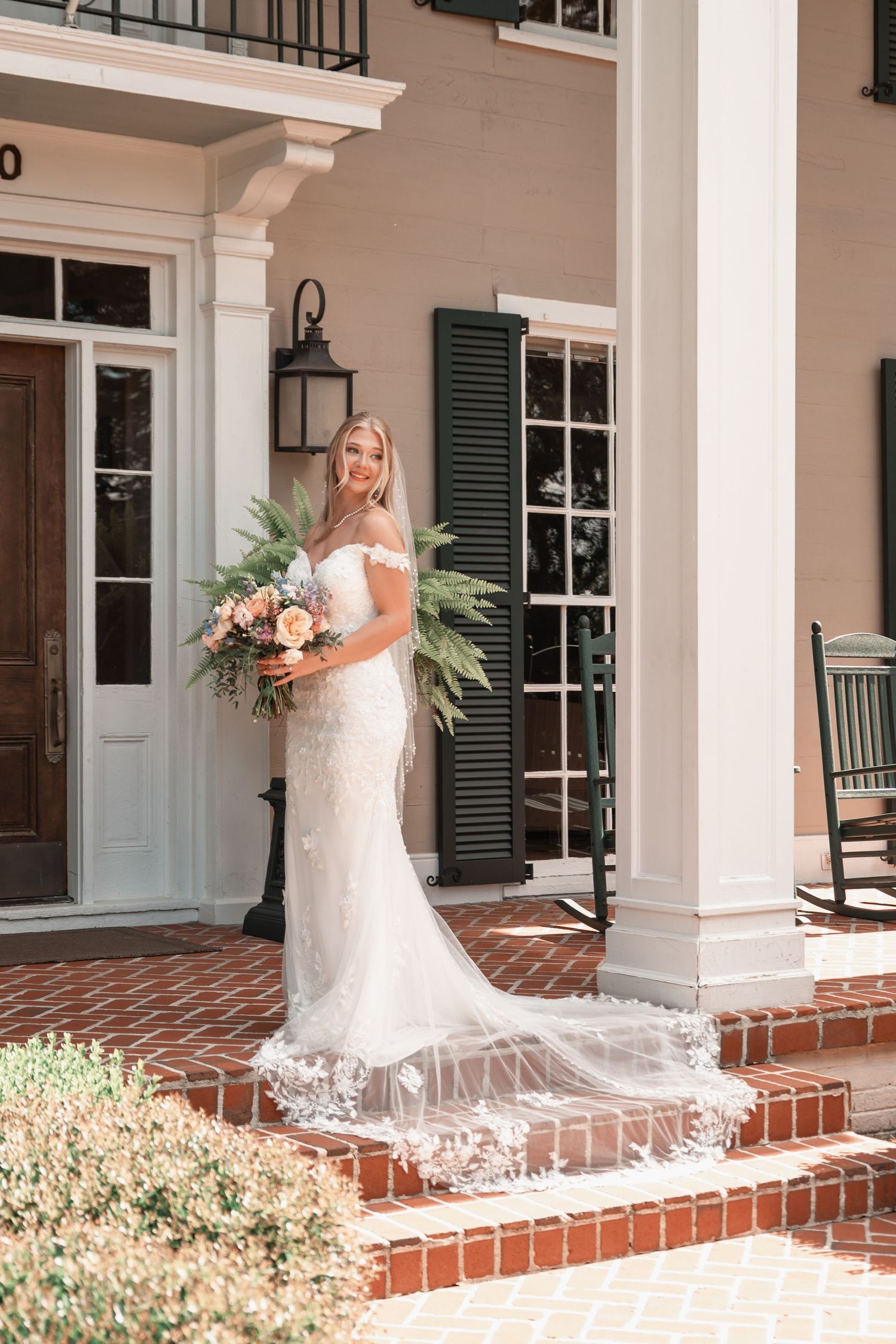 Bride in lace off-shoulder gown with floral bouquet and veil on brick steps of elegant Southern estate.