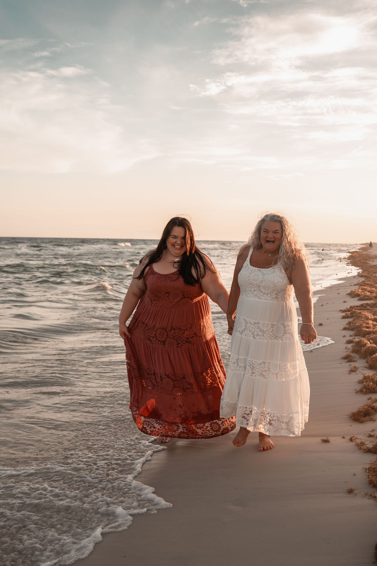 Two women in boho dresses walk hand-in-hand along a sunlit beach at sunset.