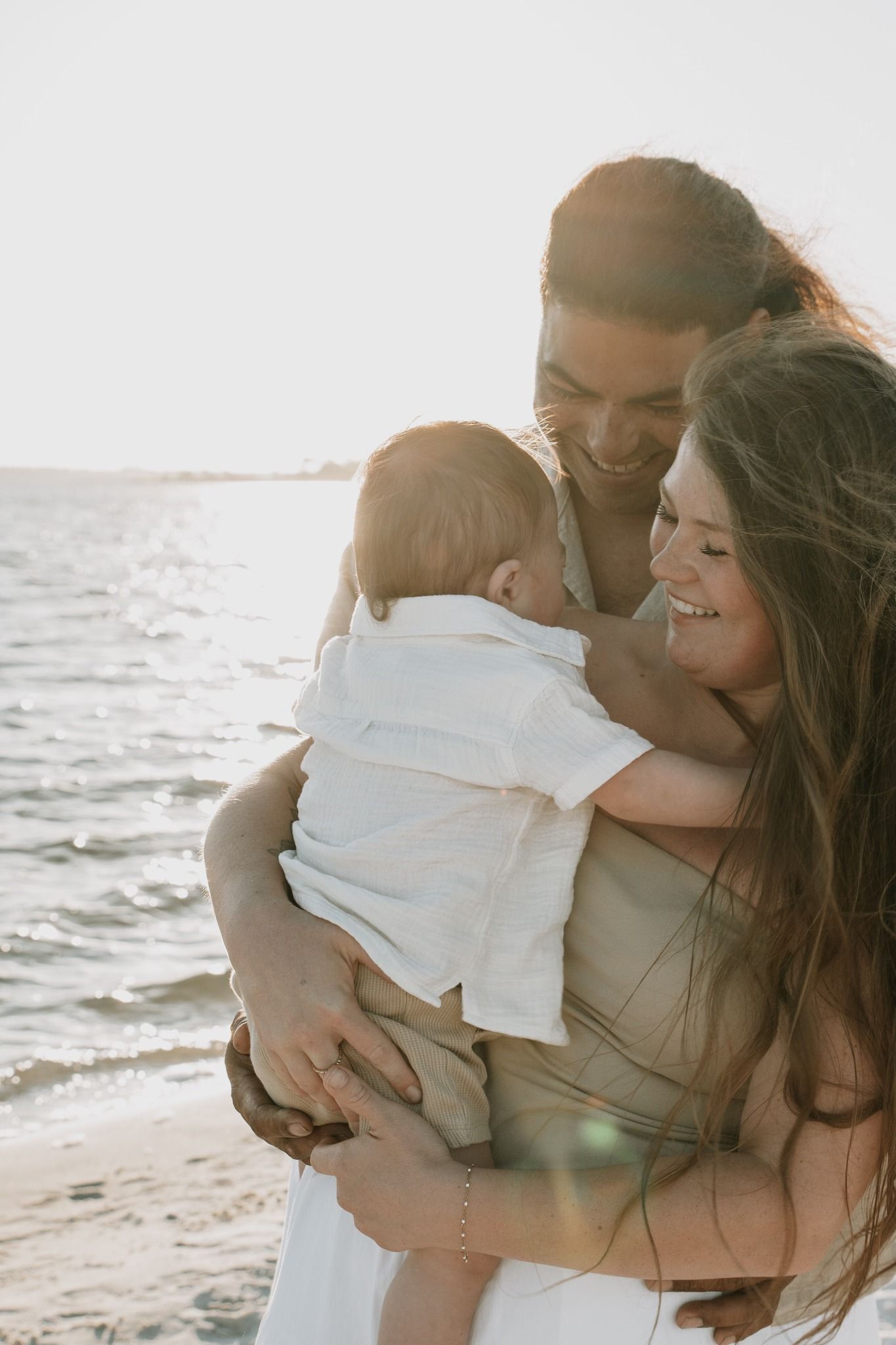 Happy young family with a baby shares a joyful embrace on a sunny beach at golden hour.