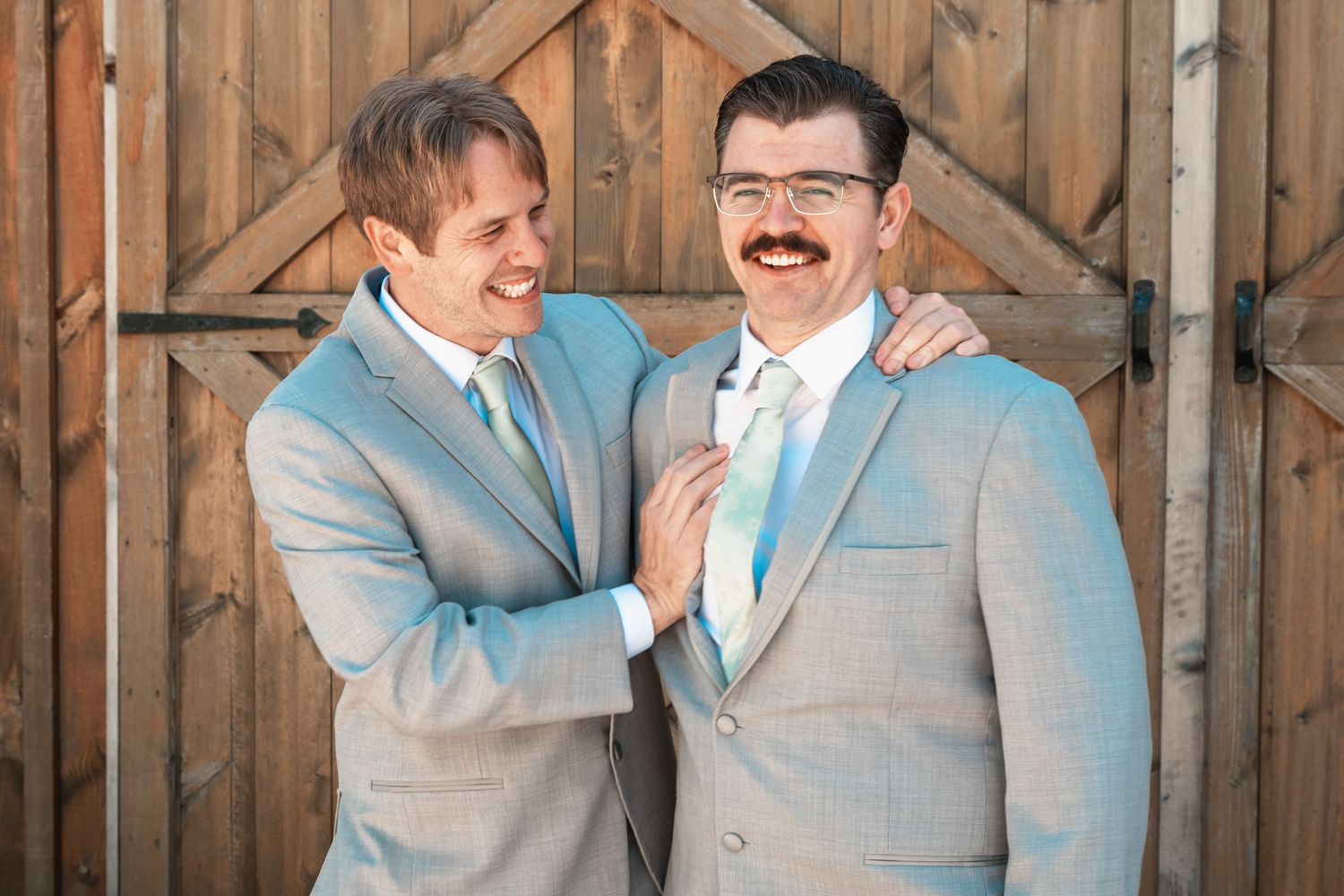 Two groomsmen in matching gray suits share a laugh in front of rustic barn doors.