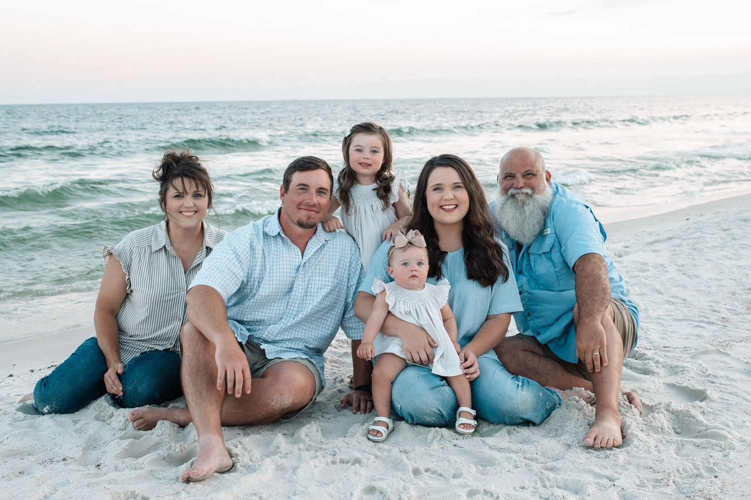 Family of six smiling together on a sandy beach with calm ocean waves in the background.
