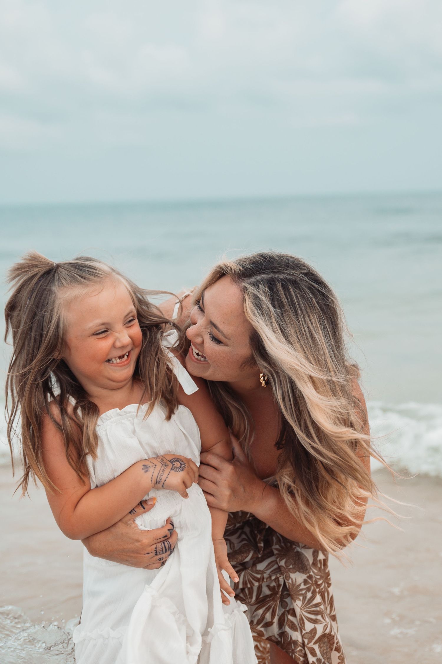 A woman and young girl with Down syndrome laugh joyfully together on a beach, sharing a tender moment by the ocean.