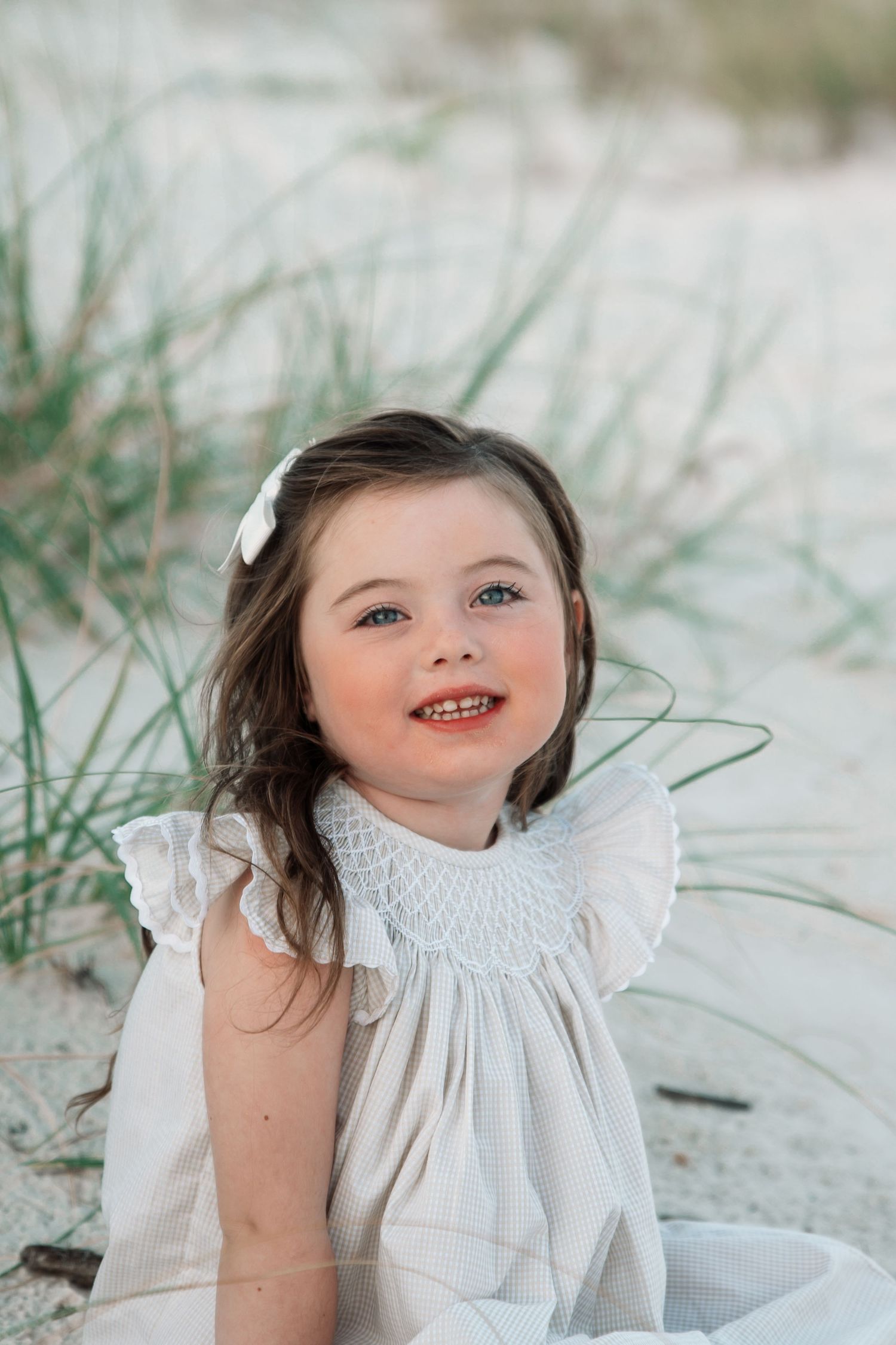 Smiling young girl with brown hair and blue eyes wearing a white smocked dress sitting on sandy beach dunes.