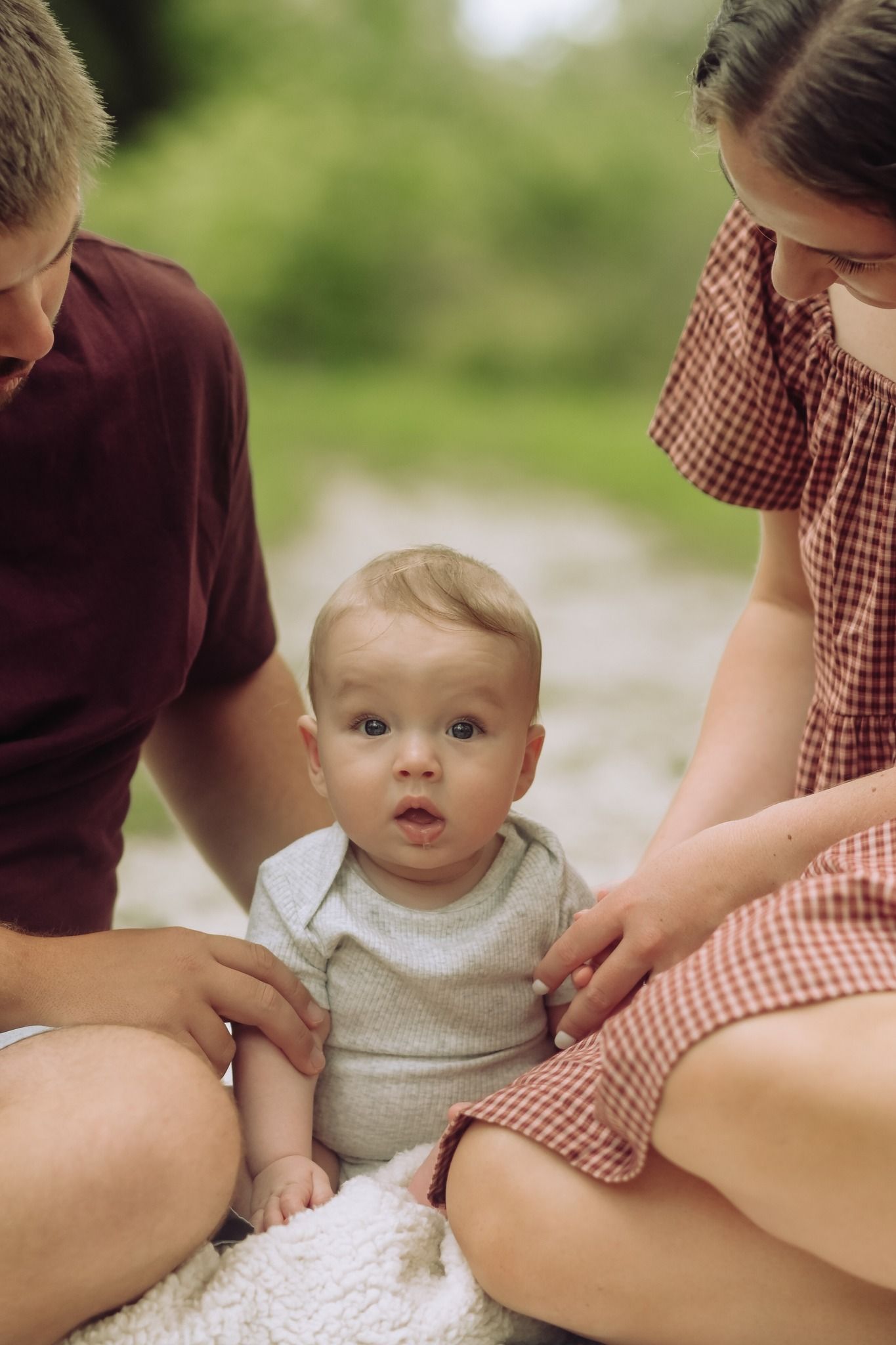 Young parents sit outdoors on a blanket with their curious wide-eyed baby between them.