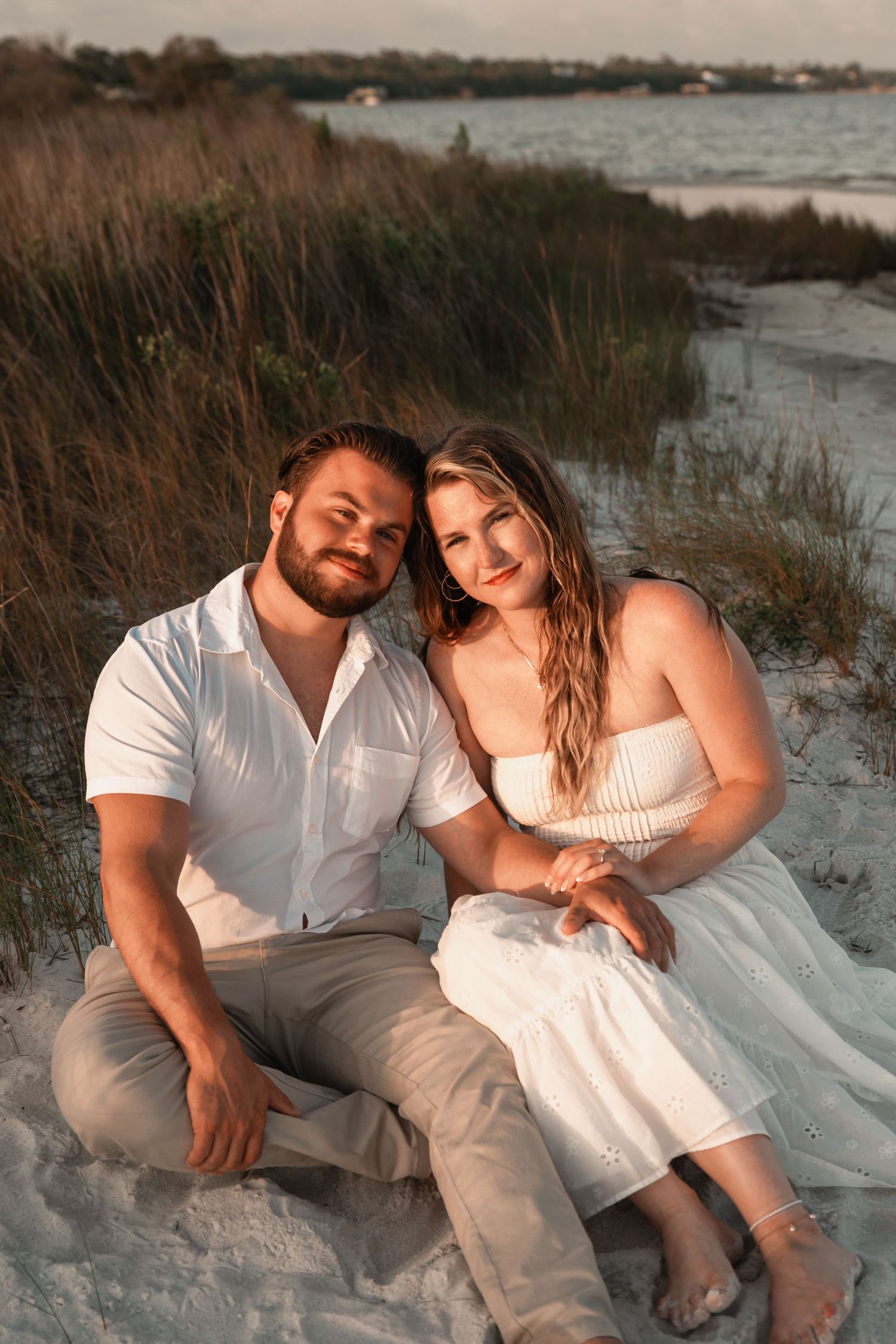 Smiling engaged couple sitting on sandy beach dunes at sunset, woman in white strapless dress.