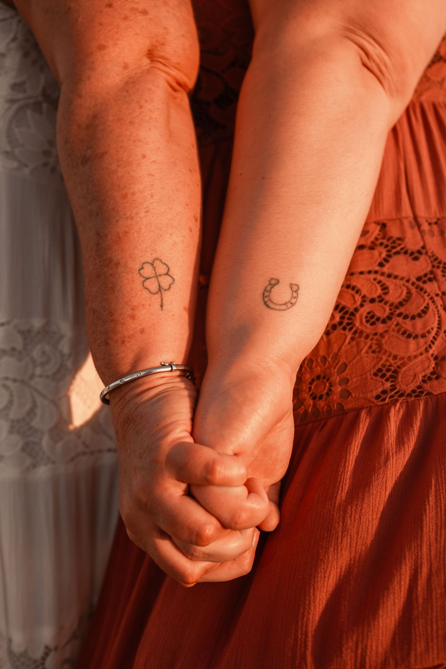 Two people holding hands showing matching lucky charm tattoos, a four-leaf clover and horseshoe, on their forearms.