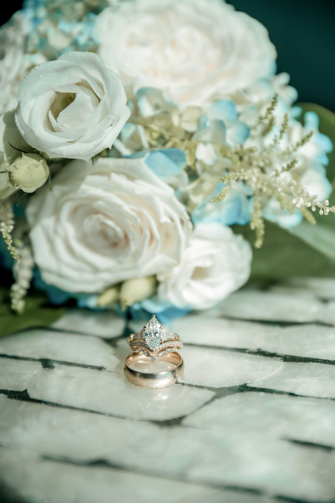 Wedding rings with diamond engagement ring rest against white and blue floral bouquet with roses on wooden surface.
