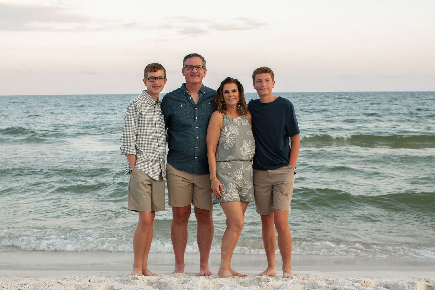 Family of four smiling together on a sandy beach at sunset with calm ocean waves in the background.
