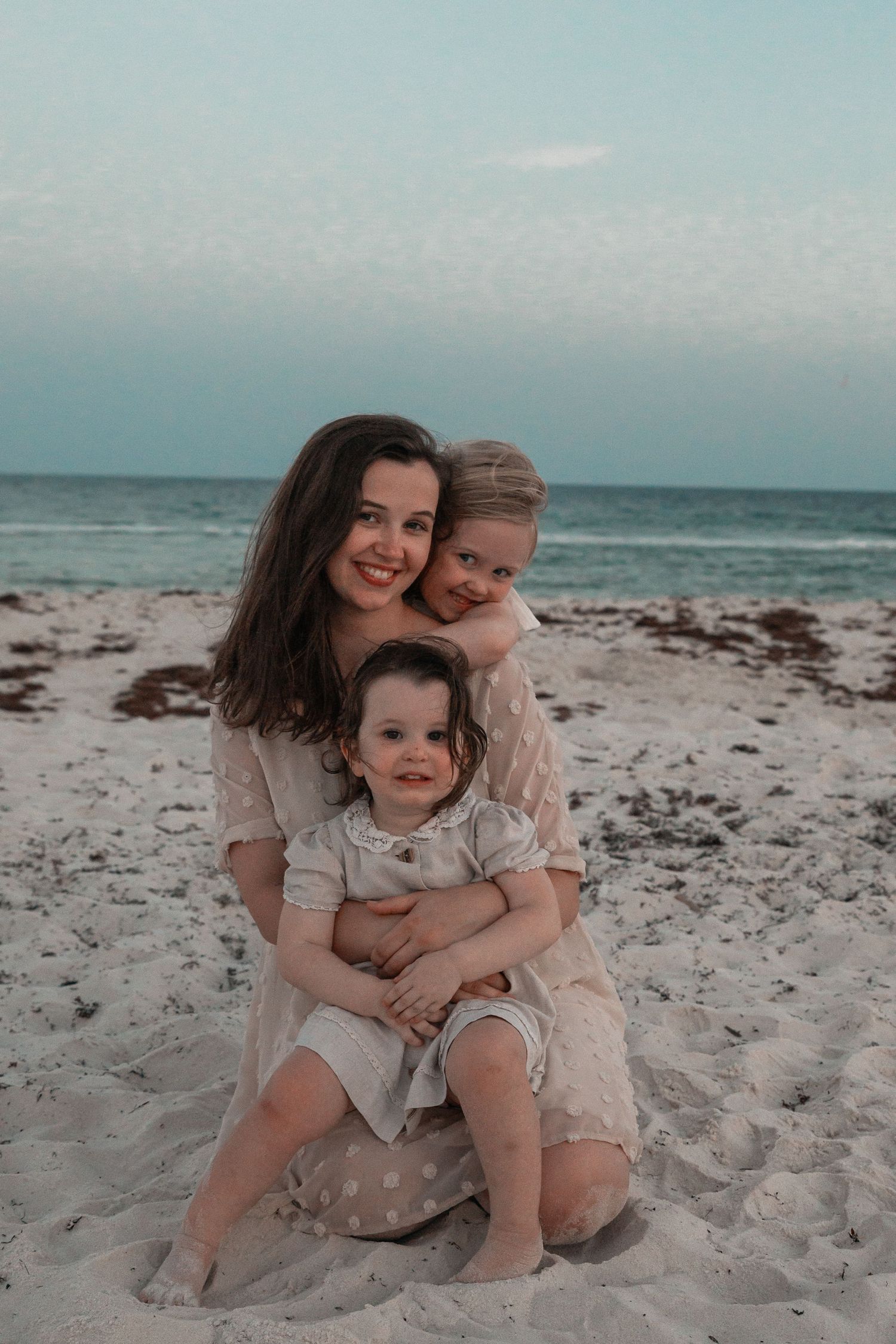 A smiling mother kneels on a white sand beach hugging two young children at sunset.