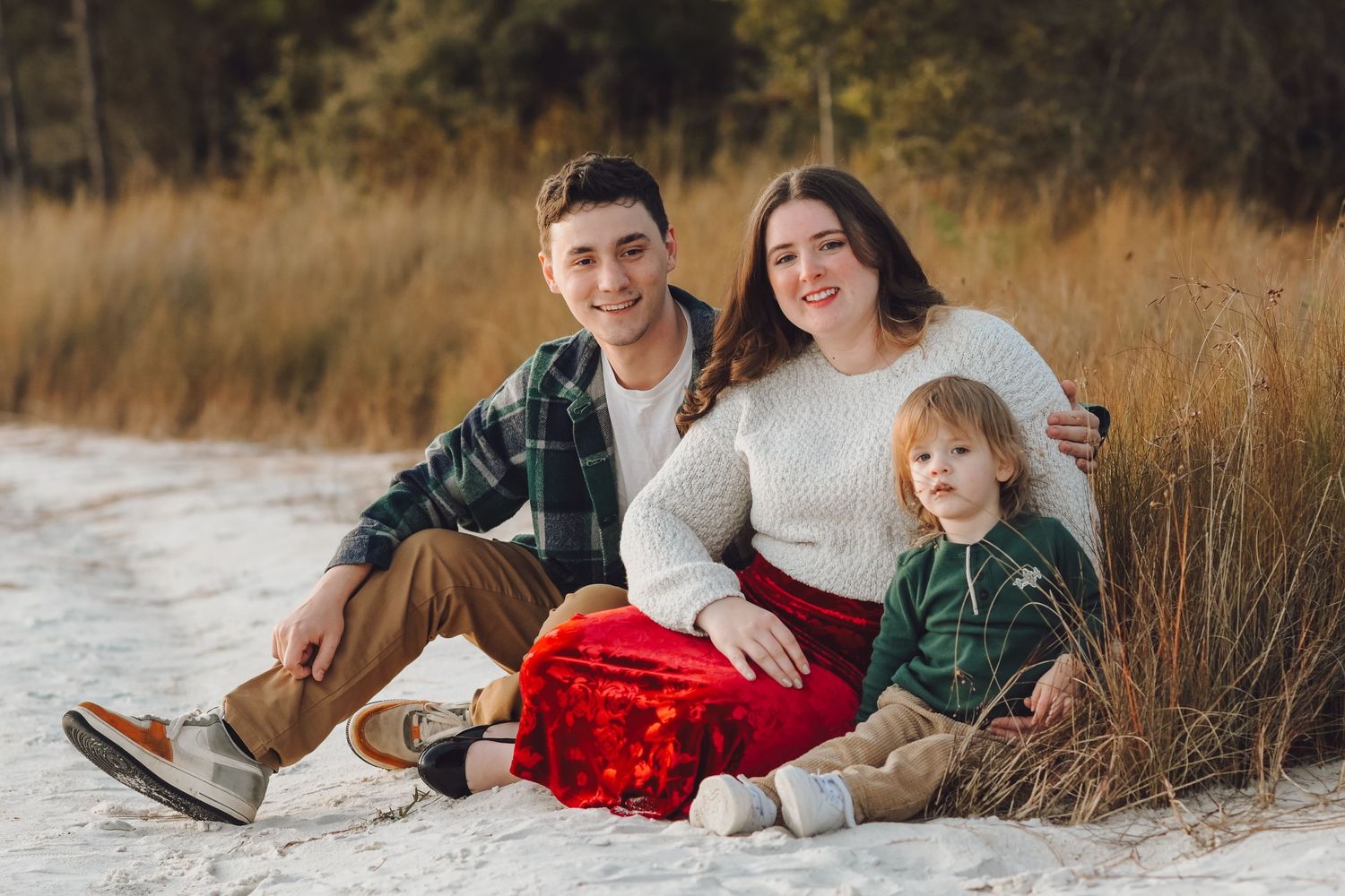 A young family of three sits together on sandy ground surrounded by golden grasses.