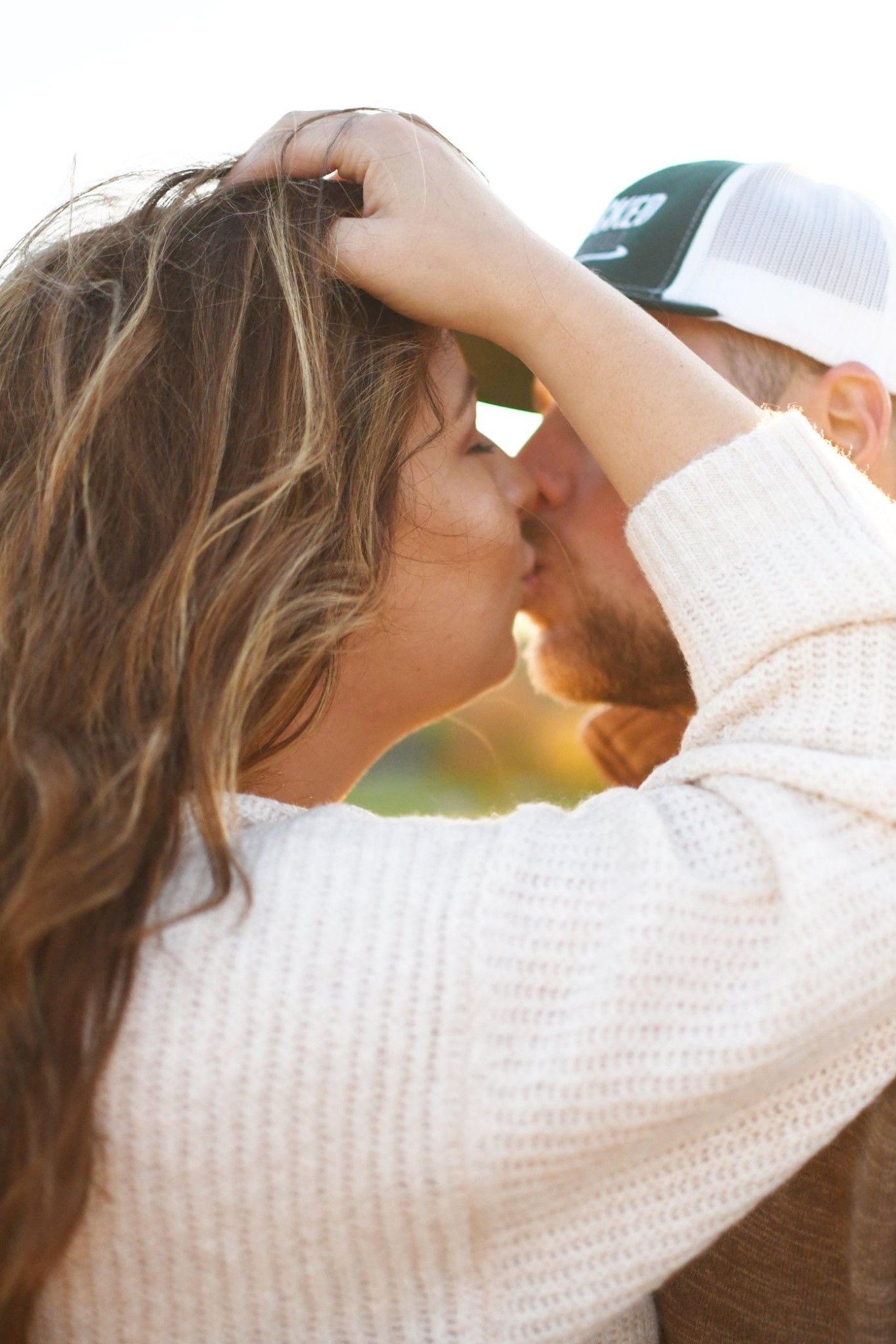 Couple sharing a close romantic kiss outdoors in golden sunset light, woman in white sweater holding mans cap.