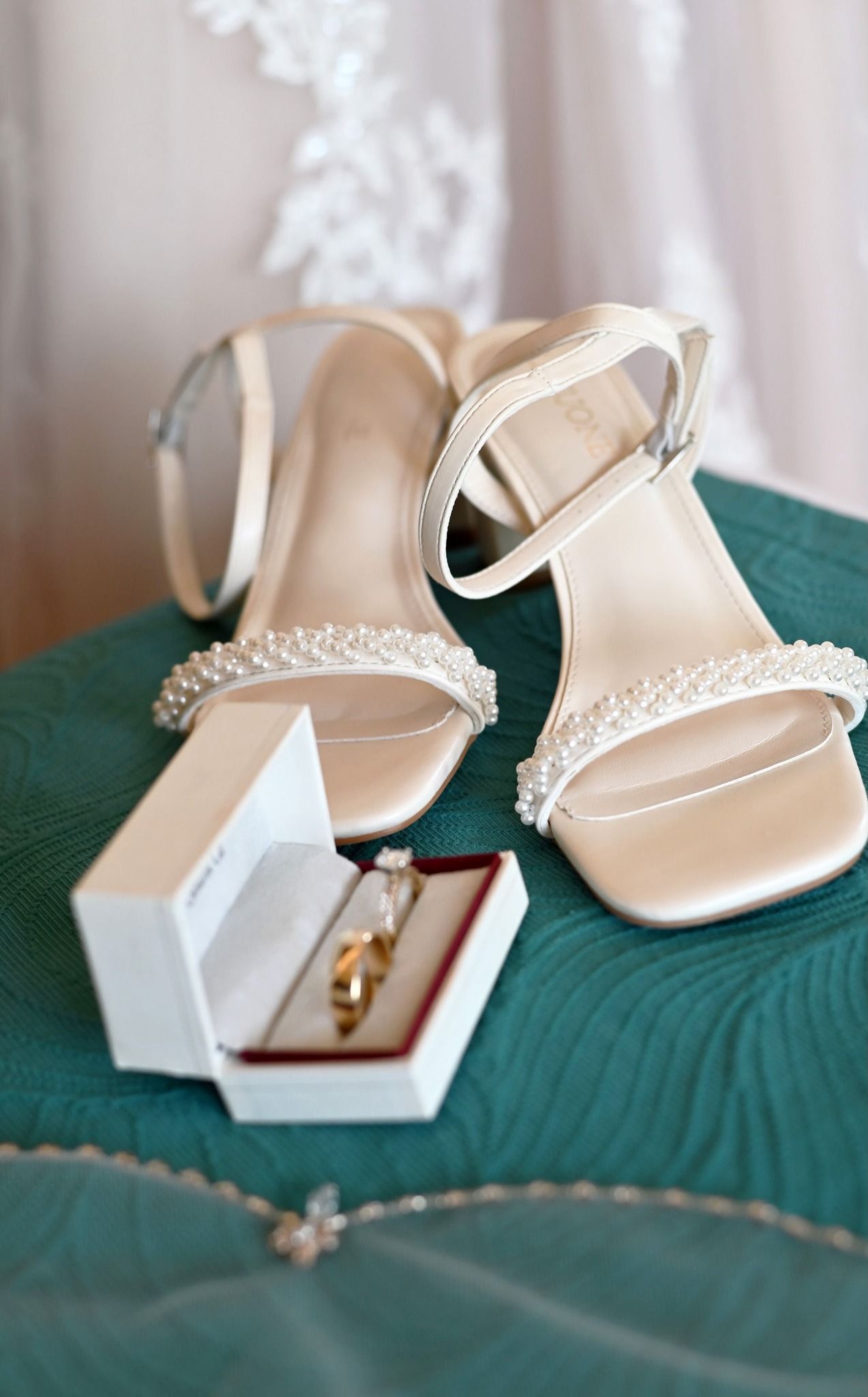 White pearl-strap bridal heels displayed with gold wedding rings in red box on teal surface.